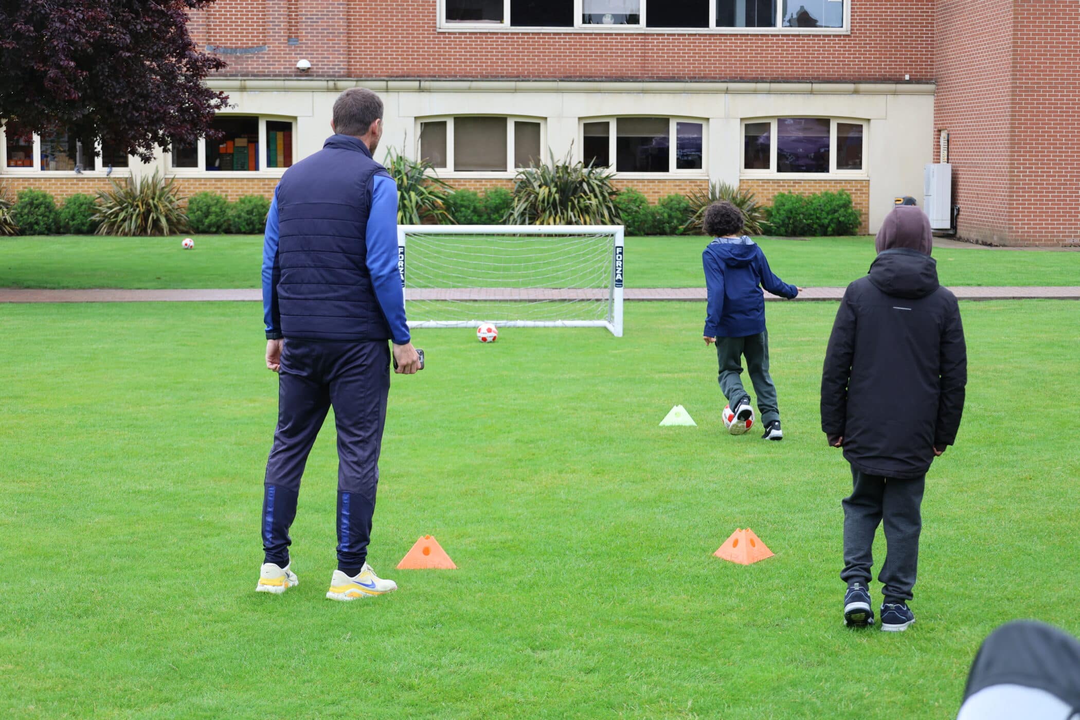 A coach and two children are on a grassy field, practising football during Open Events. One child is about to kick a ball towards a small goal, while the other stands nearby. Orange cones mark the drill, with a brick building in the background.