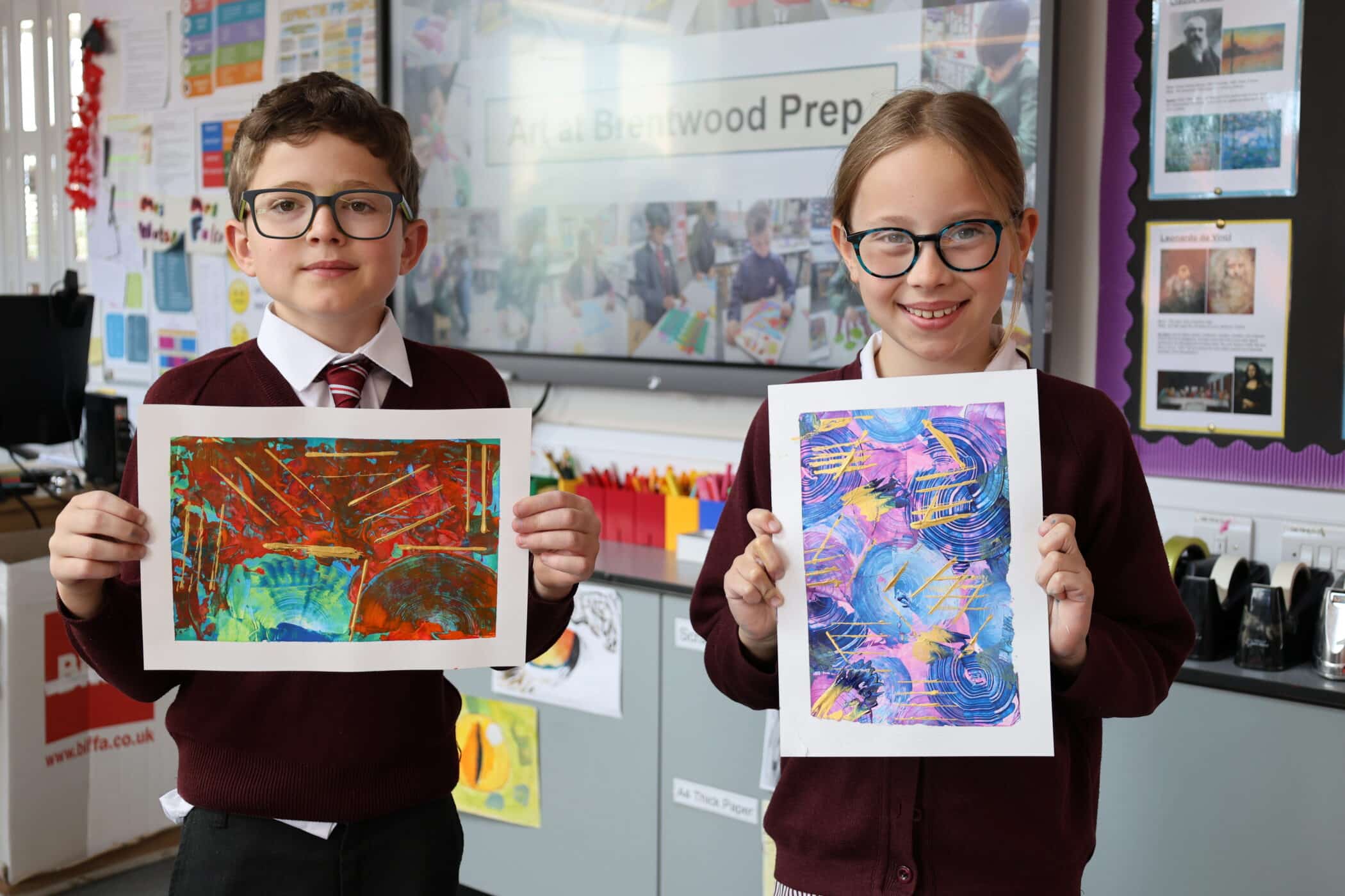 Two schoolchildren in uniforms stand in a classroom, proudly displaying colourful abstract art. Bright decorations and an