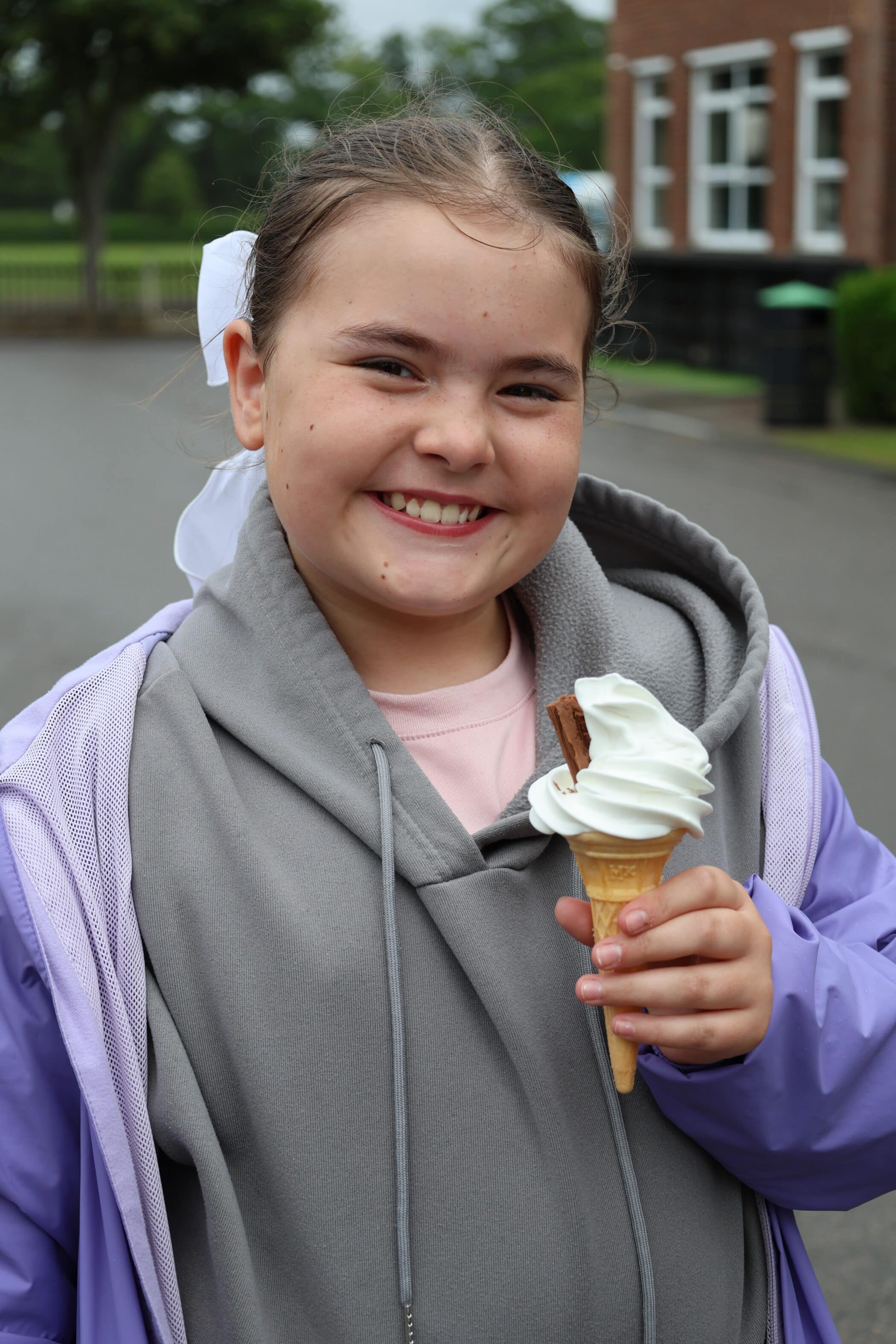 A smiling young girl in a purple jacket holds a vanilla ice cream cone with a chocolate flake, enjoying an Open Events day outside on a paved area with trees and a brick building in the background.