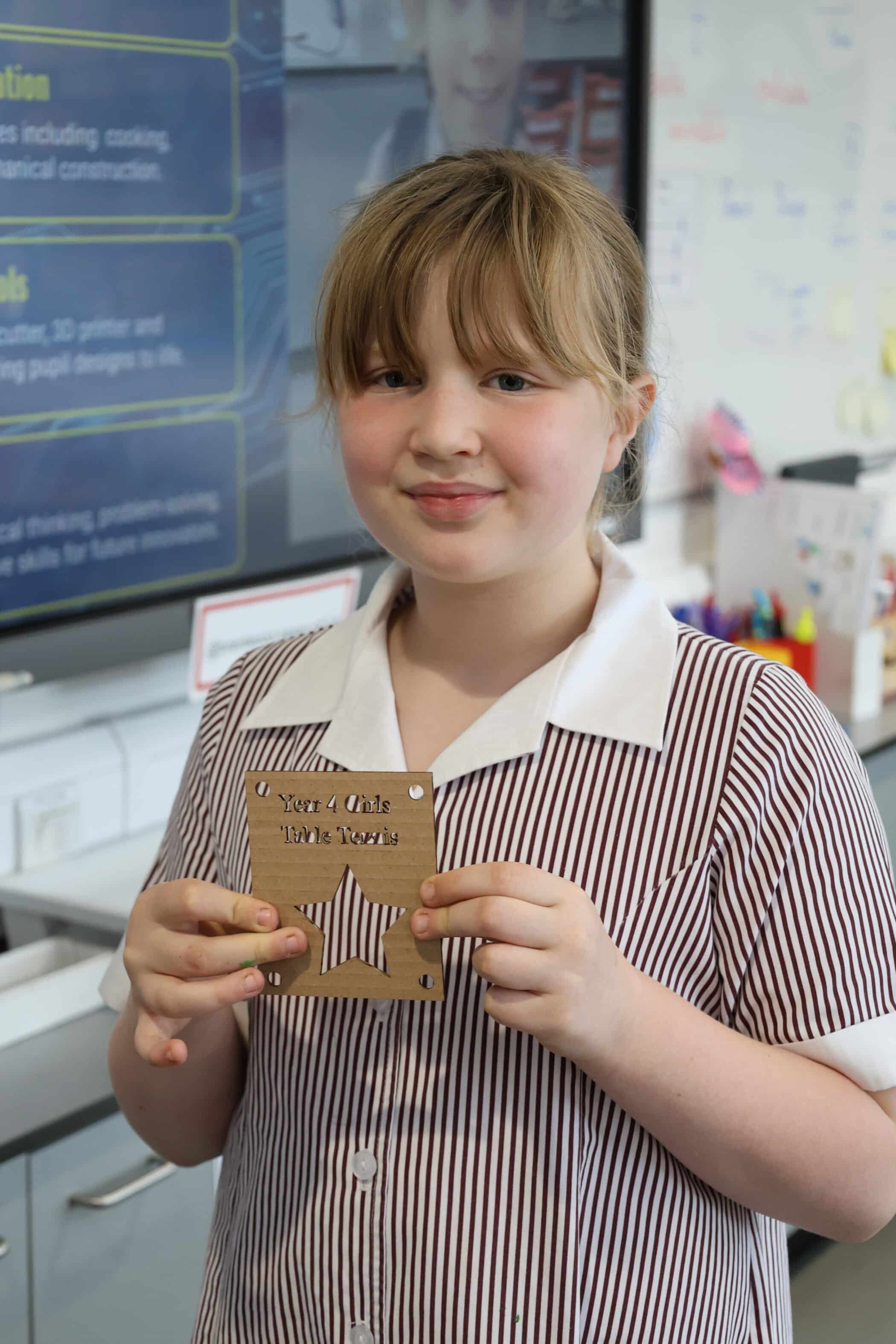 A young pupil in a striped uniform holds a wooden award plaque reading Year 4 Girls' Maths Award in a classroom with a digital screen and whiteboard, proudly representing her school at Open Events.