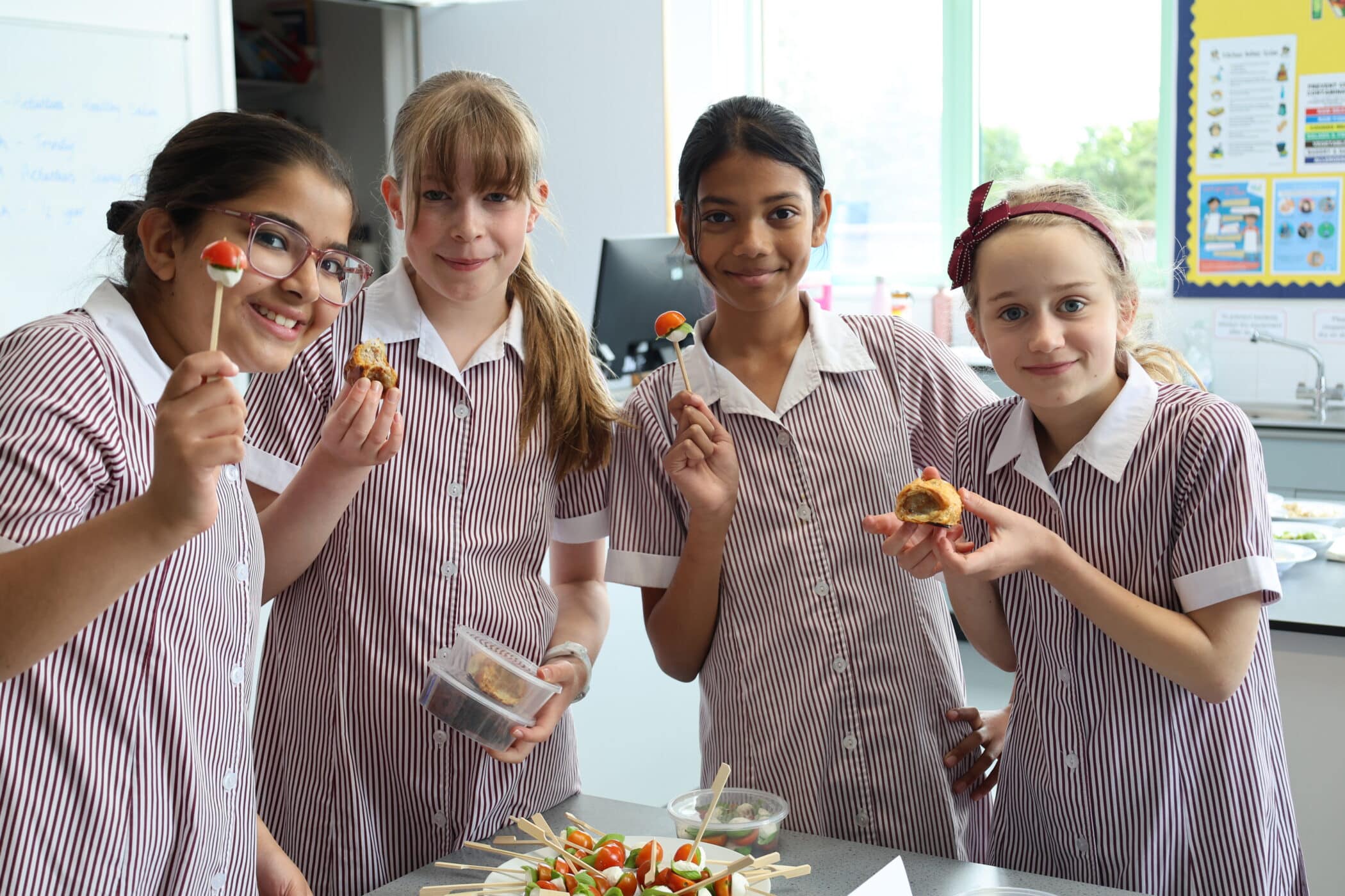 Four schoolgirls in striped uniforms smile and hold skewered snacks in a classroom or kitchen setting during one of the Open Events; food and ingredients are spread out on the table in front of them.