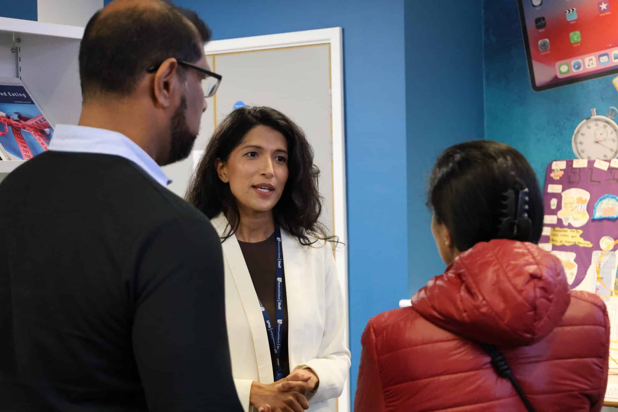 A woman in a white blazer speaks to two people, one in a black jumper and the other in a red jacket, during Open Events in a brightly lit room with blue walls and colourful posters.
