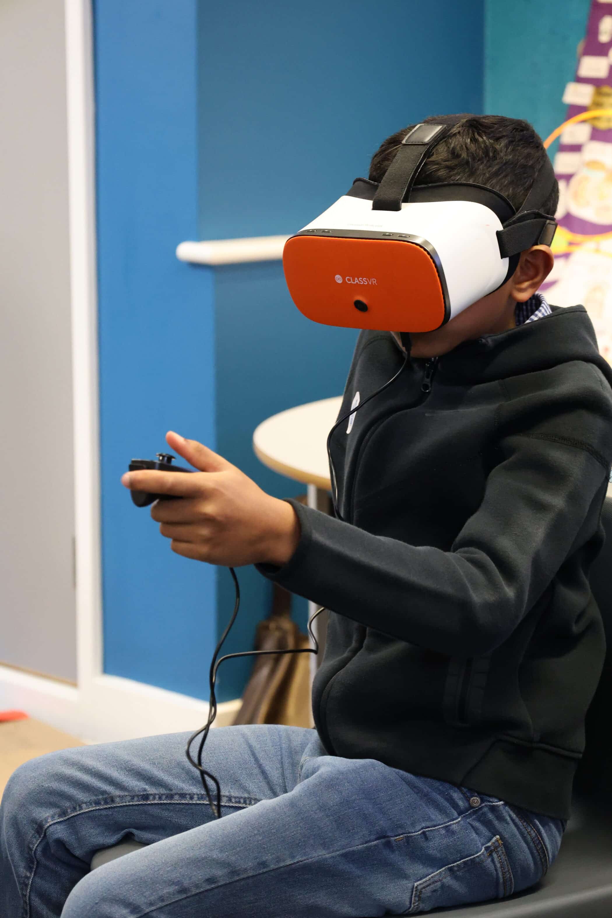 A boy wearing a black hoodie and blue jeans sits in a chair using a virtual reality headset and holding a controller, exploring Open Events, with a blue wall and table in the background.