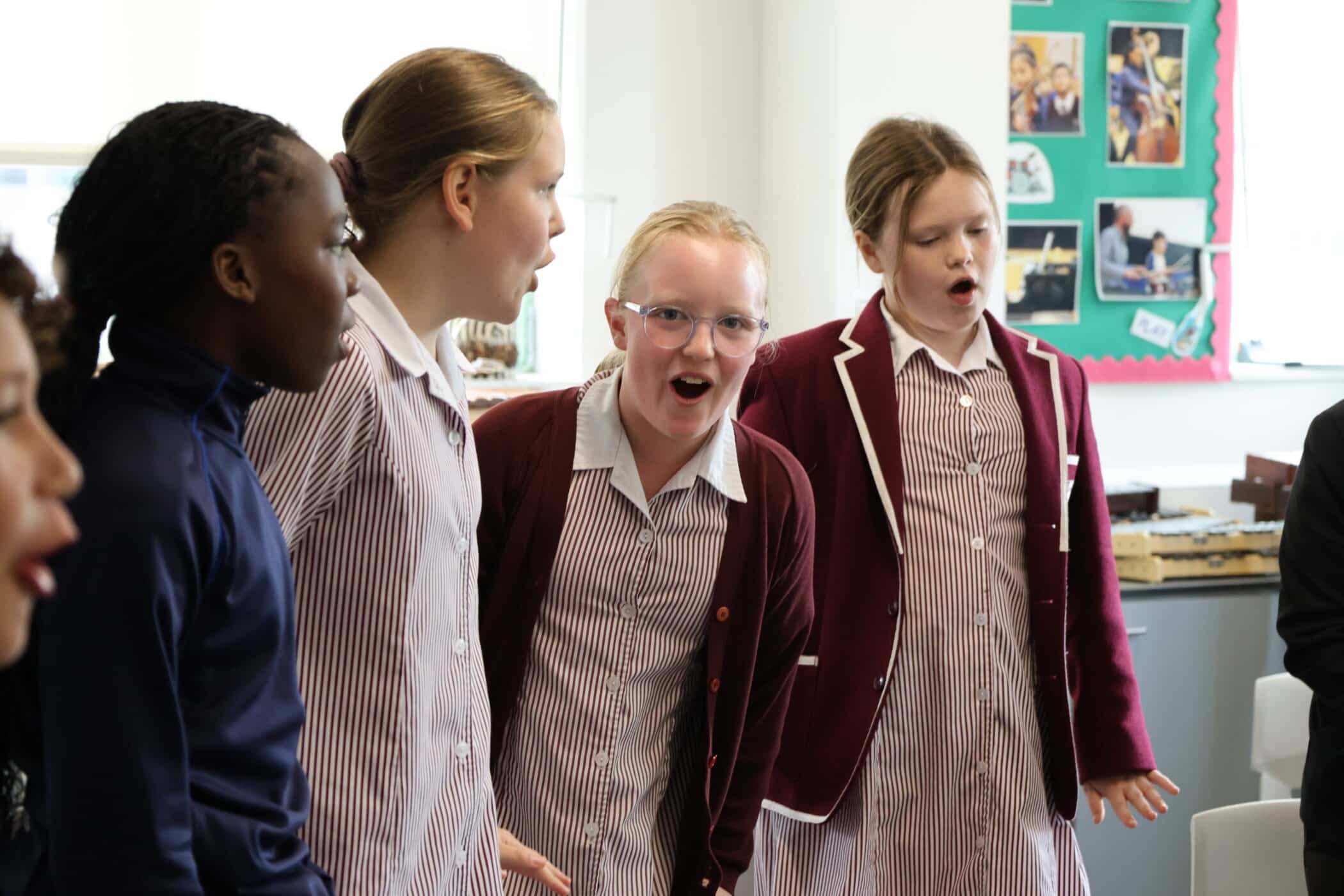 Four schoolgirls in uniforms stand closely together, enthusiastically singing or speaking with expressive faces—capturing the lively spirit often seen during Open Events. A green noticeboard with photos is visible in the background.