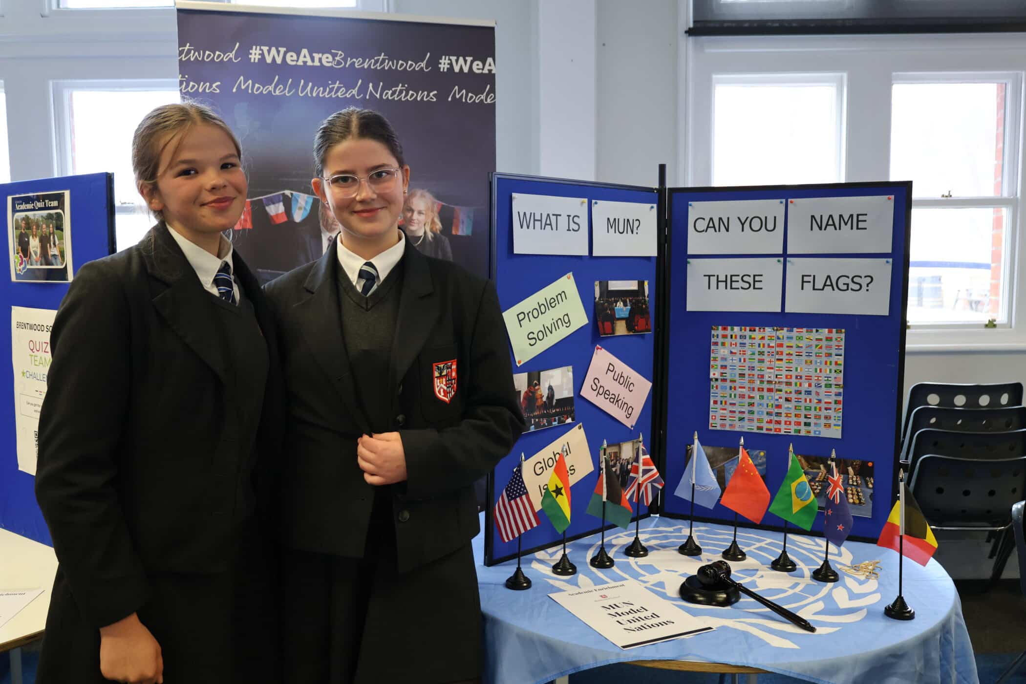 Two students in school uniforms stand by a Model United Nations display at an Open Events day, featuring small flags, world maps, and signs asking “Can you name these flags?” and “What is MUN?” in a classroom setting.