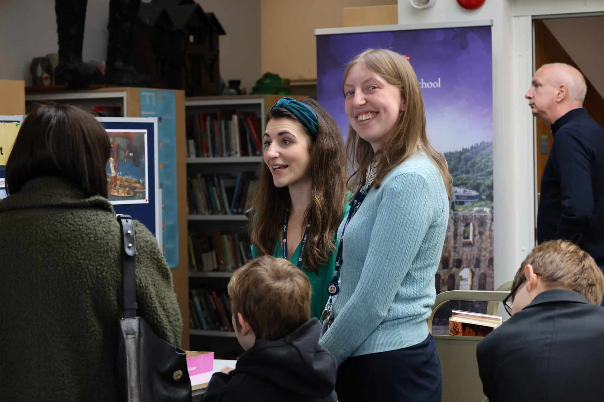 Two women stand smiling and talking to visitors at an Open Events gathering near bookshelves; children and adults are also present, creating a lively, engaging atmosphere.