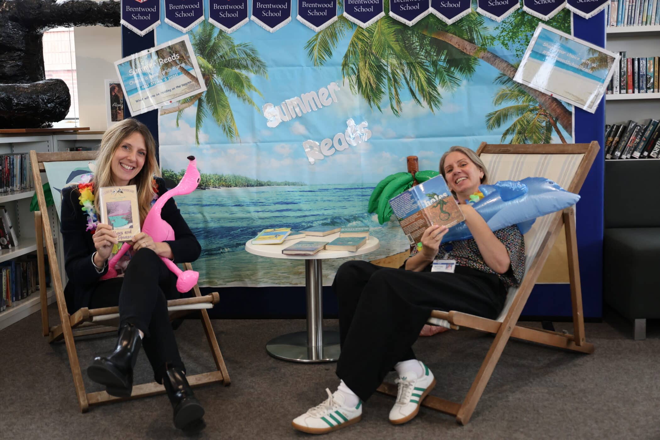 Two women sit on deckchairs in a library decorated with a tropical beach backdrop. Surrounded by books and beach toys, they enjoy Summer Reads as part of the library’s Open Events, with featured titles displayed on a small table.