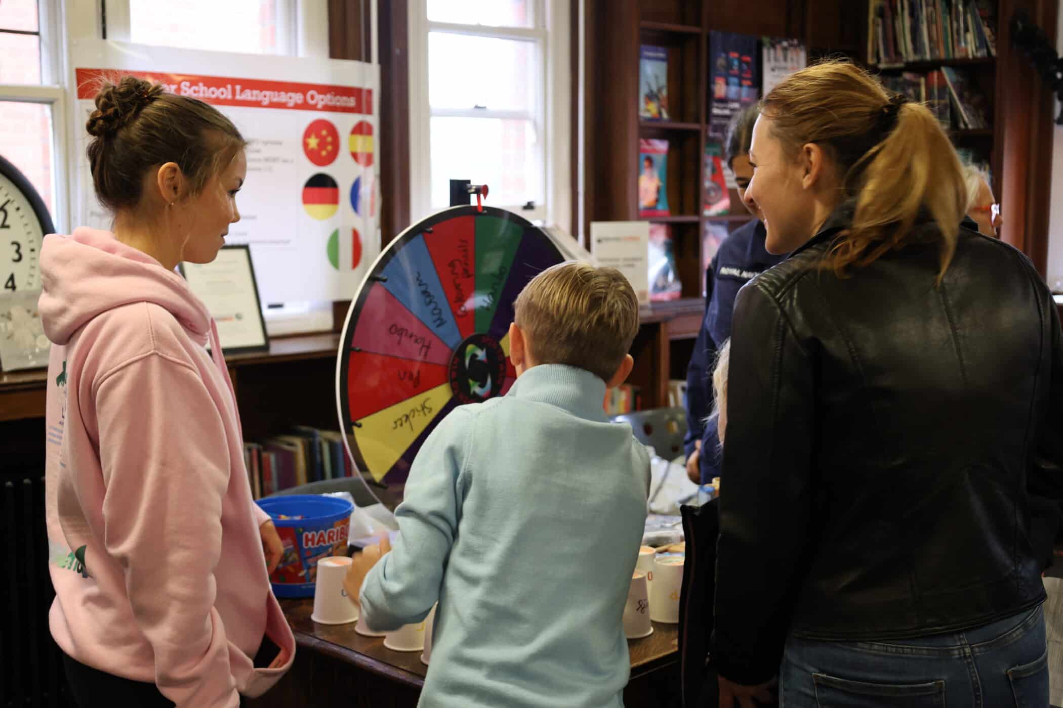 Three people stand around a table playing a spin-the-wheel game during Open Events in a room lined with bookshelves. A colourful prize wheel is the focus, surrounded by cups and sweets on the table nearby.