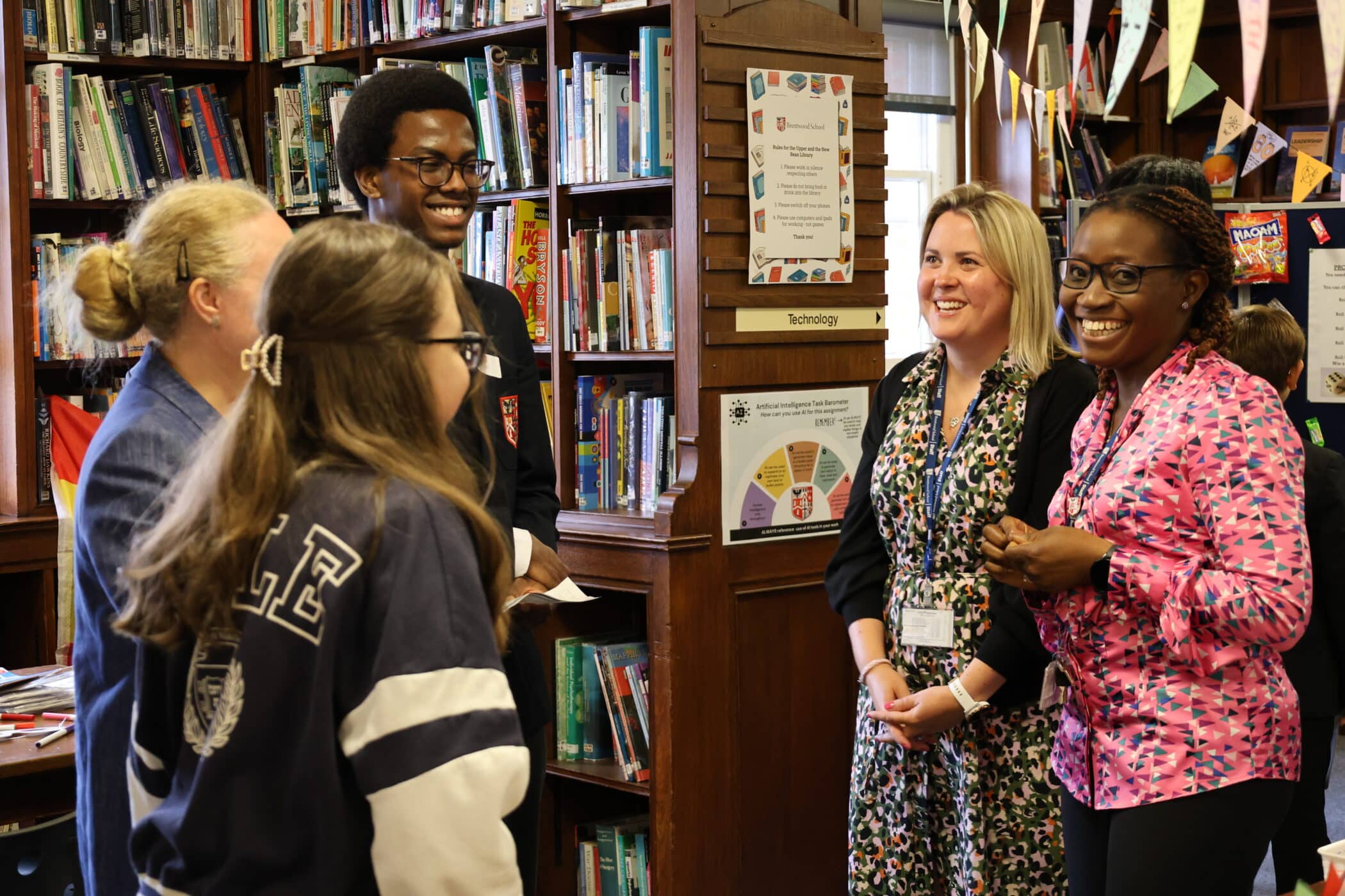 Five people, including students and adults, stand and chat in a library filled with bookshelves, colourful bunting, and educational posters during Open Events, all smiling and engaged in friendly conversation.