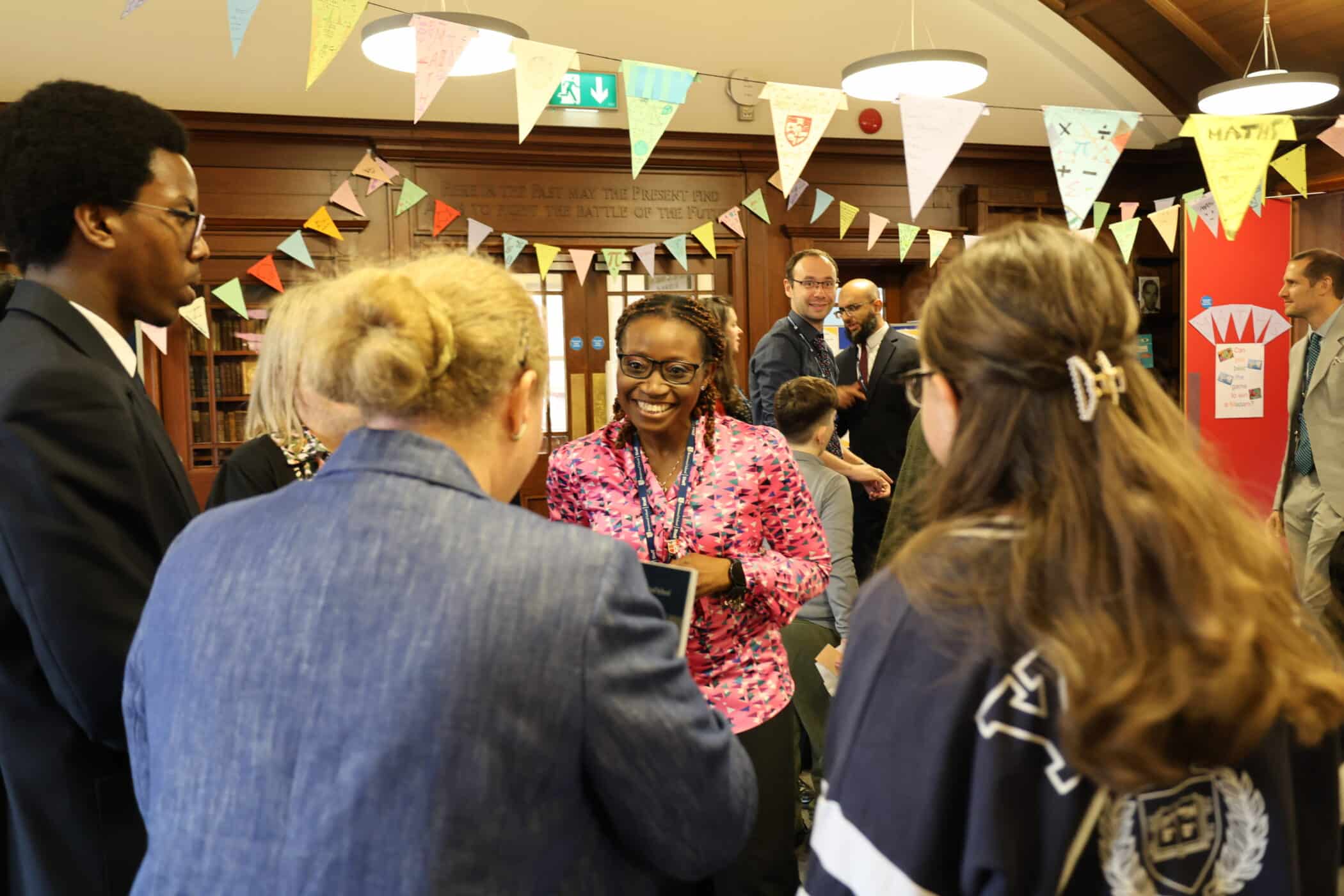 A group of people converse and smile at an Open Event decorated with colourful bunting. A woman in a pink patterned shirt is speaking, whilst others listen and interact in a friendly, lively atmosphere.