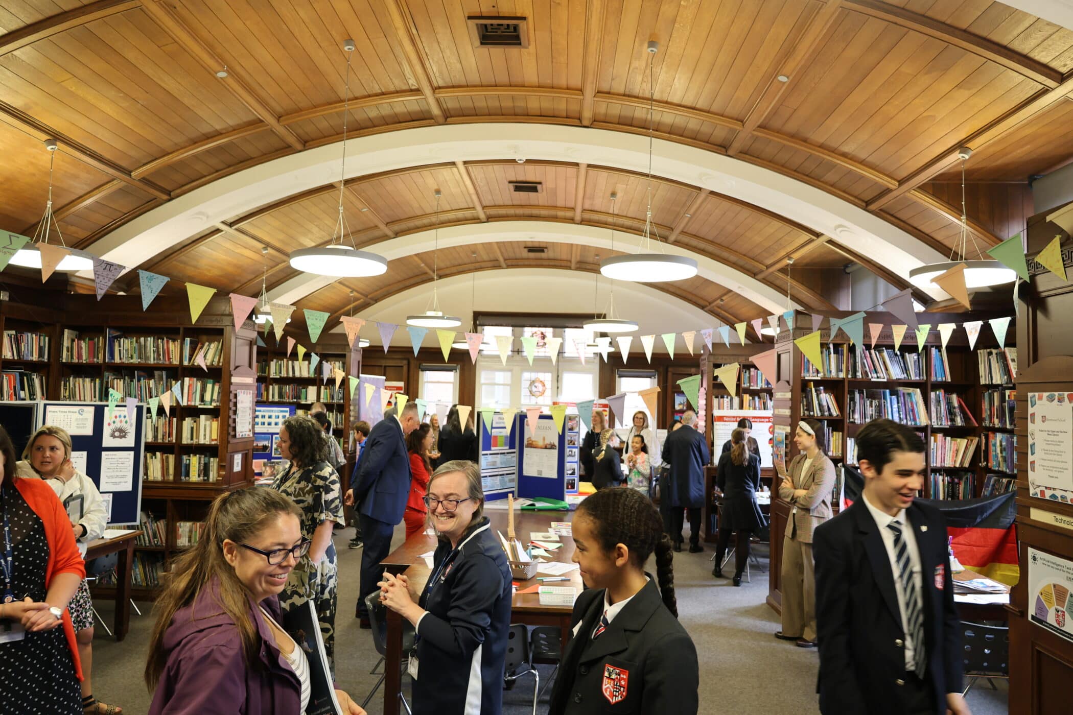 A busy library with a wooden ceiling, decorated with bunting for Open Events. People in business attire and students in uniforms are talking and viewing colourful display boards set up throughout the room. Shelves of books line the walls.