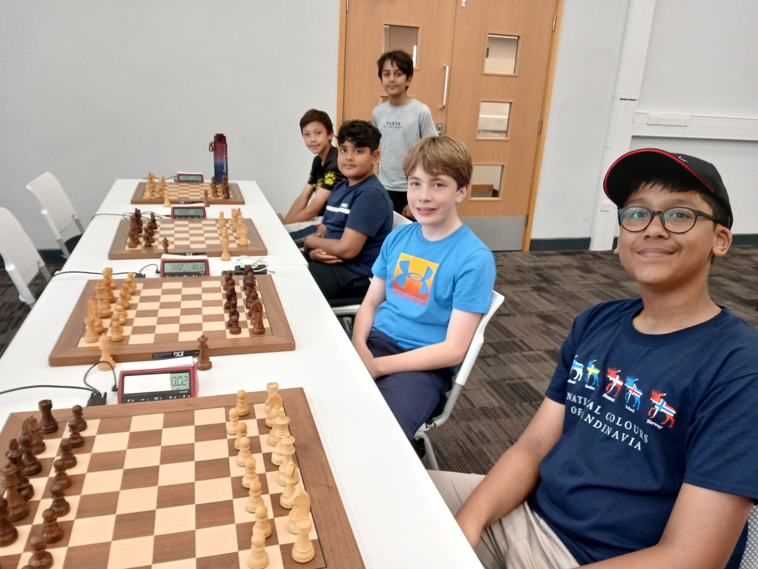Five boys are seated at a long table set up for a chess tournament, with chessboards and clocks in front of them. They are smiling, and the room has white walls and a light wooden door in the background.