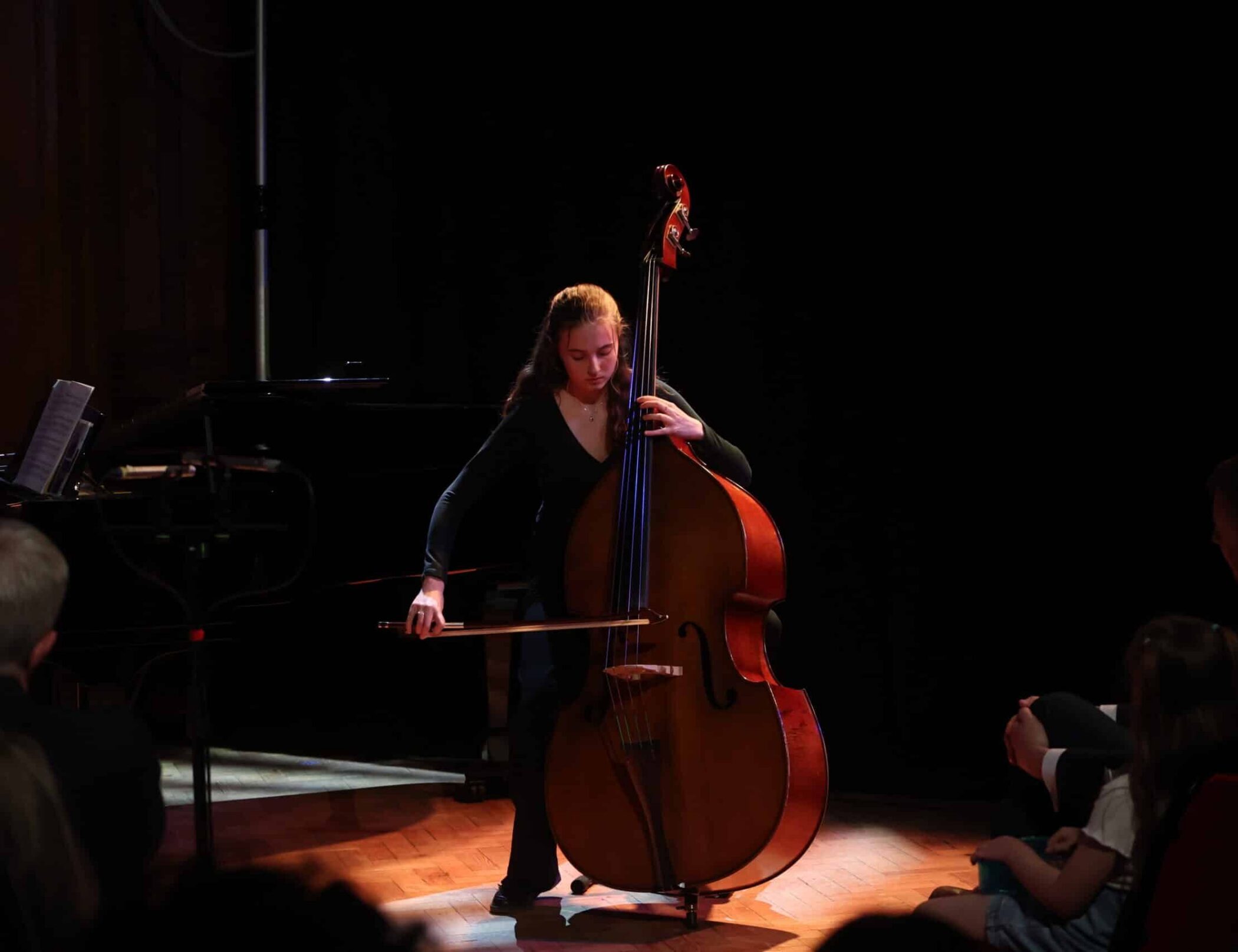 A young woman from the Specialist Music Programme plays a double bass with a bow on a dimly lit stage, whilst an audience watches. She stands in a spotlight, focused on her performance.