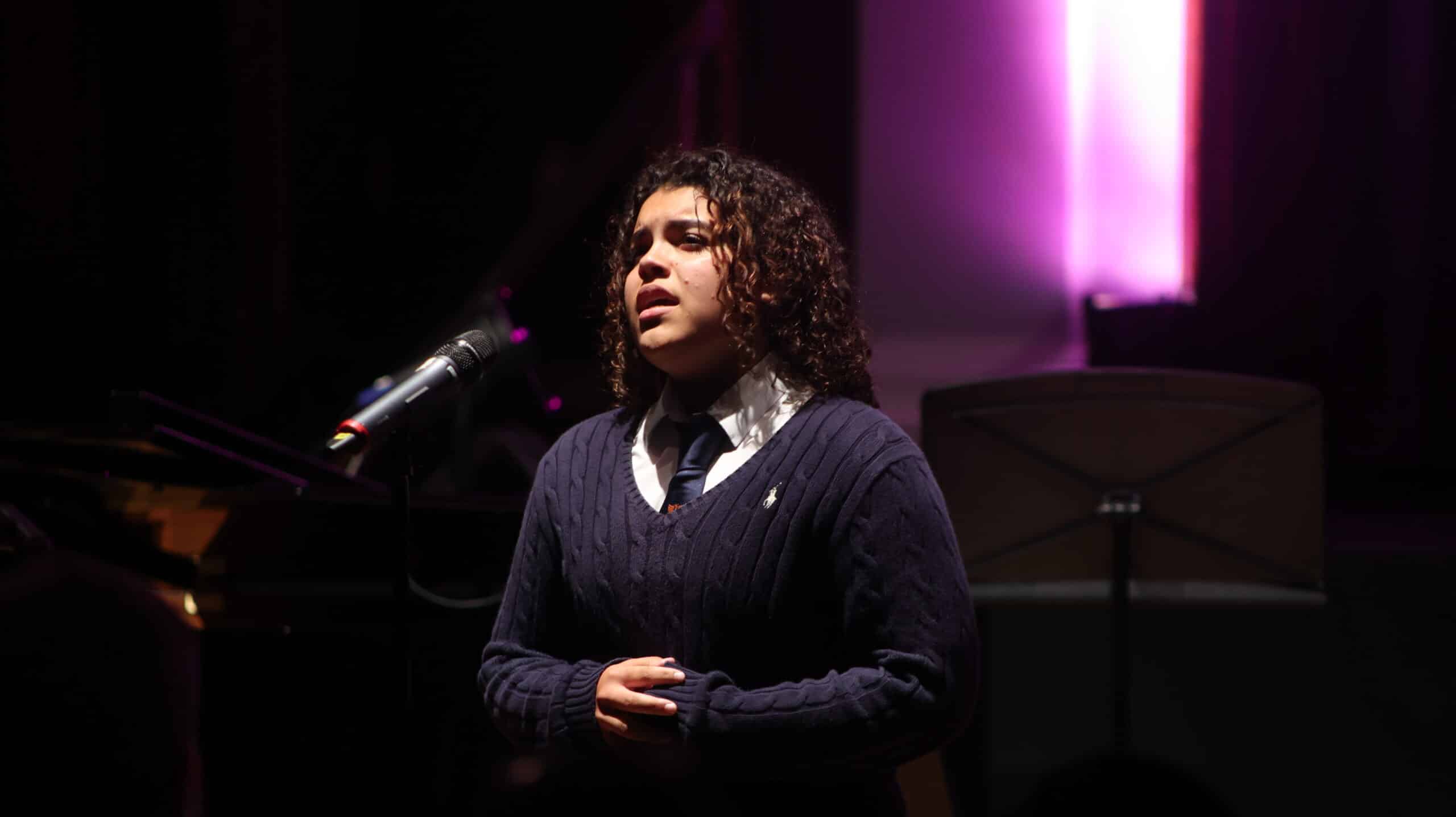 A person with curly hair sings into a microphone on stage, wearing a navy jumper and tie, under dramatic purple lighting in the background, as part of the Specialist Music Programme.