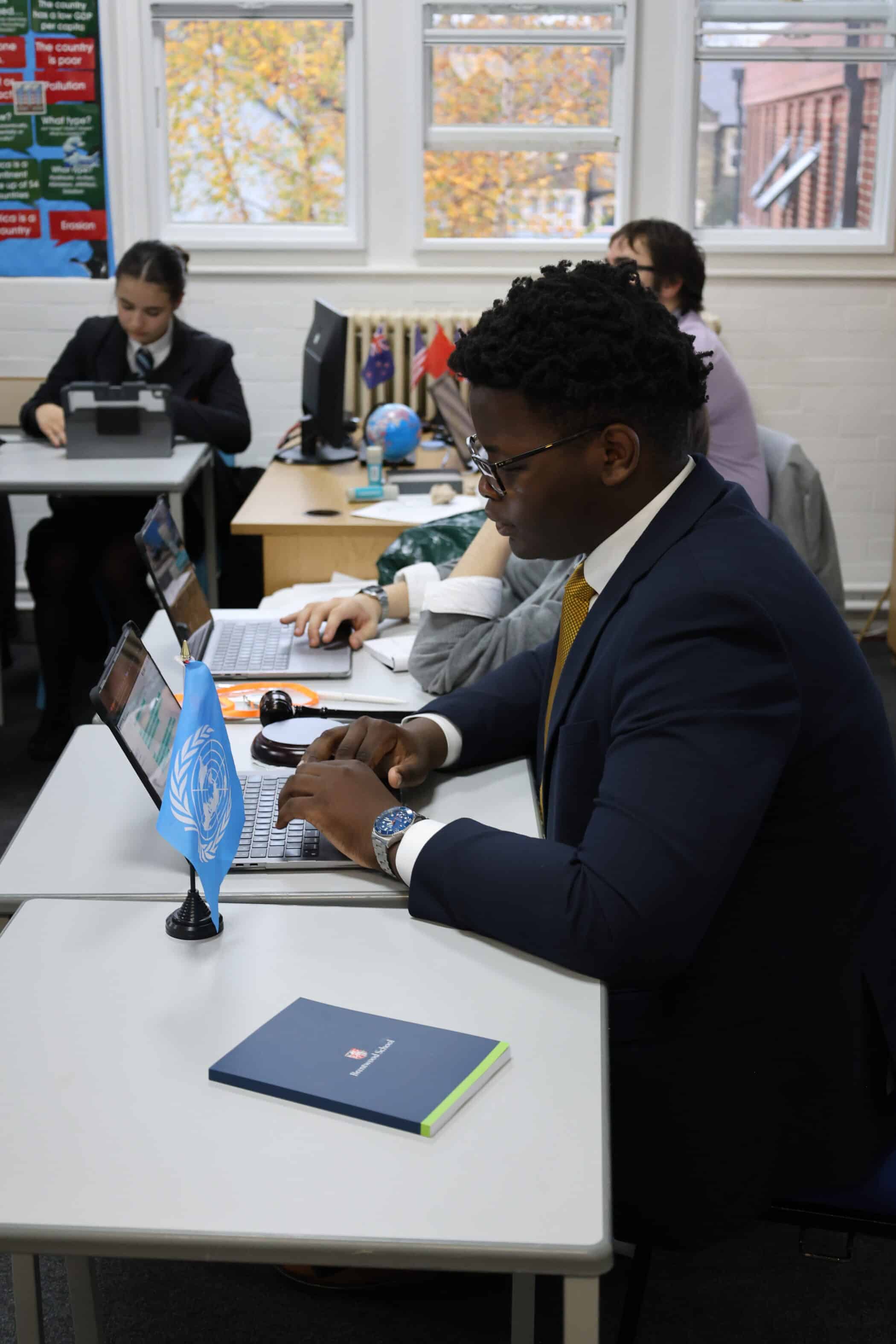 A person in a suit types on a laptop at a desk with a small UN flag, whilst others work at computers in a classroom with large windows and autumn trees outside.
