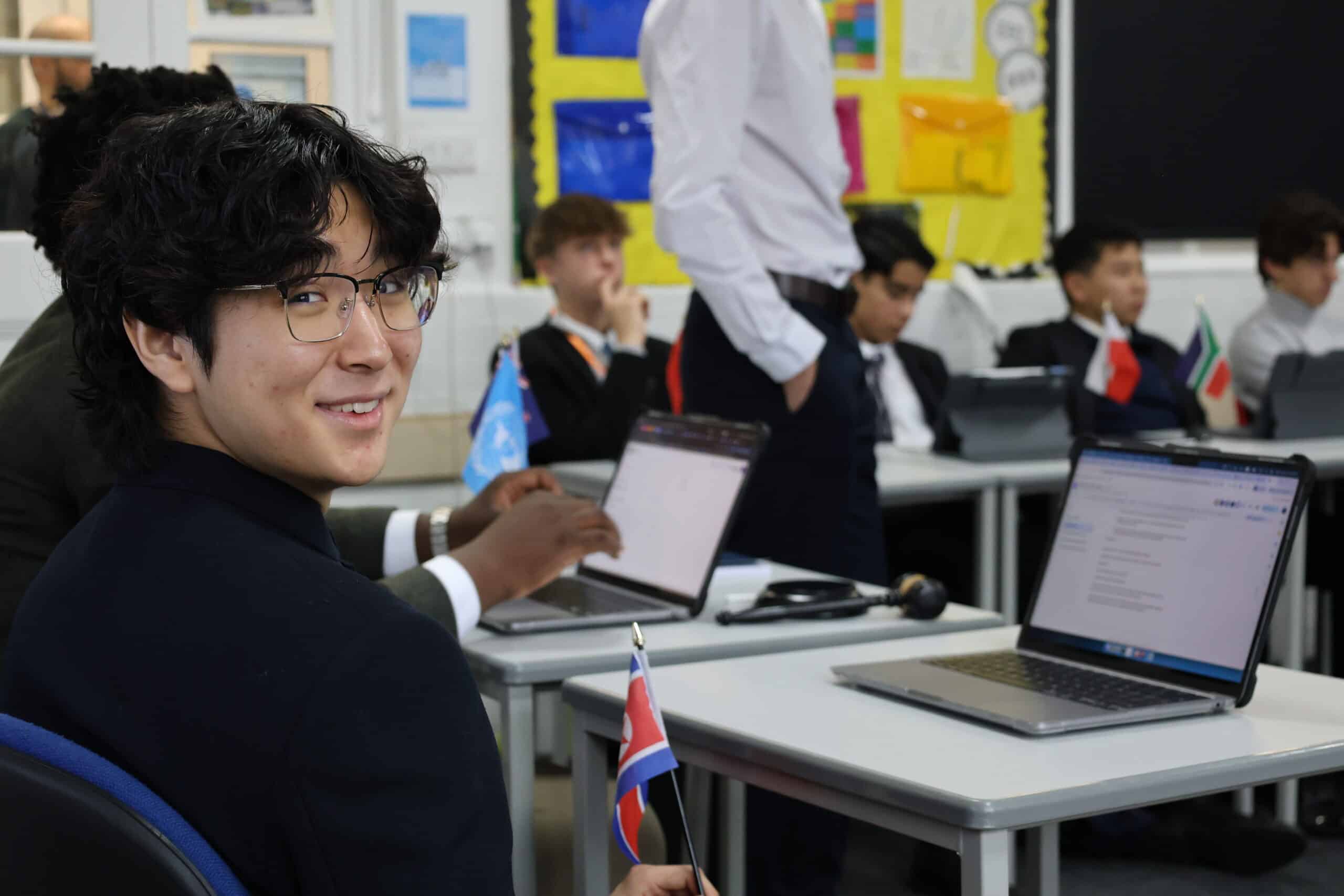 A student with glasses smiles at the camera whilst holding a small flag in a classroom, capturing the vibrant atmosphere of how Sixth Forms prepare students for university. Several other students sit at desks with laptops, and there are more flags and colourful posters in the background.