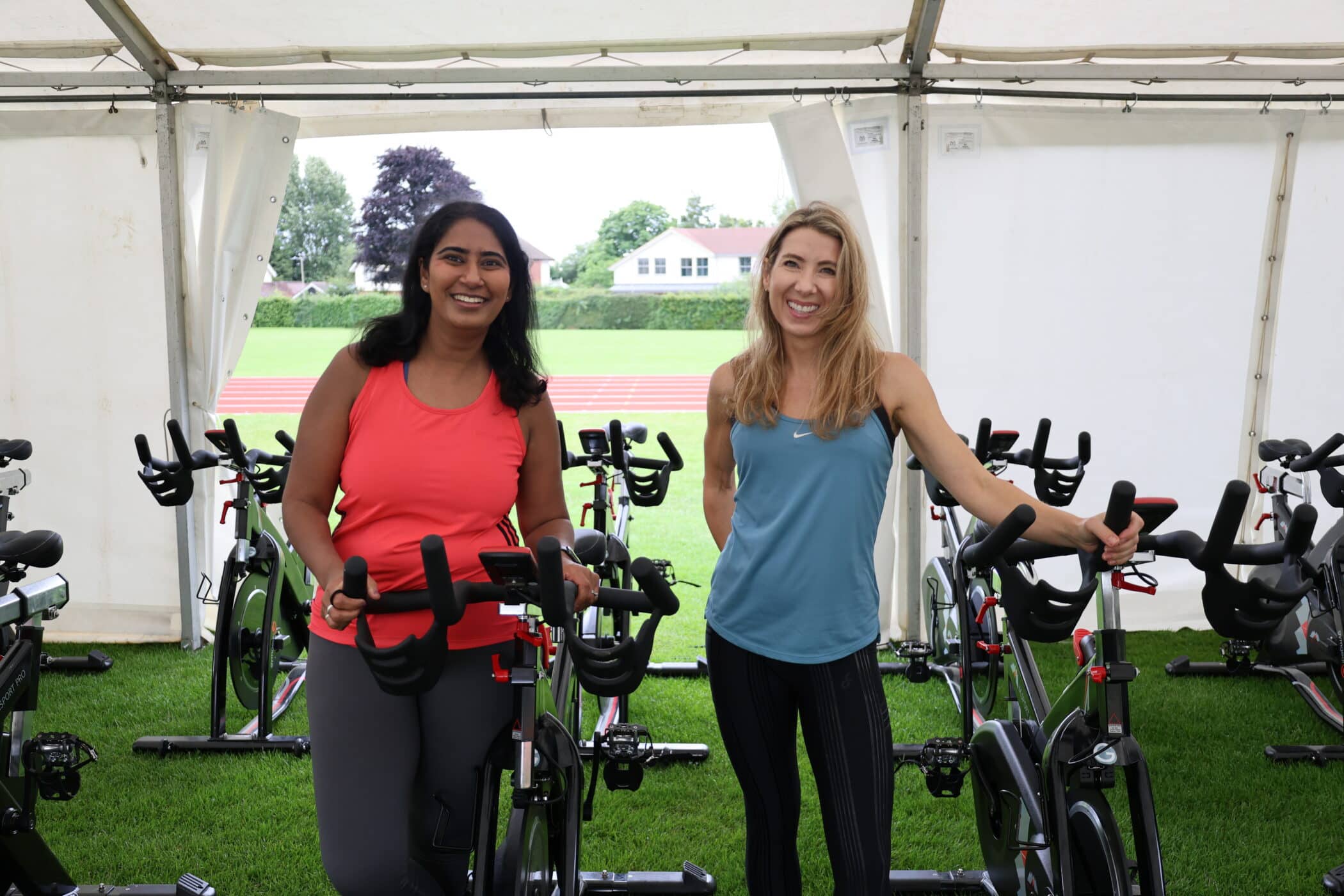 Two women smiling and standing beside stationary bikes in a marquee set up on a grassy field during a Brentwood School Foundation event, with more exercise bikes and a running track visible in the background.