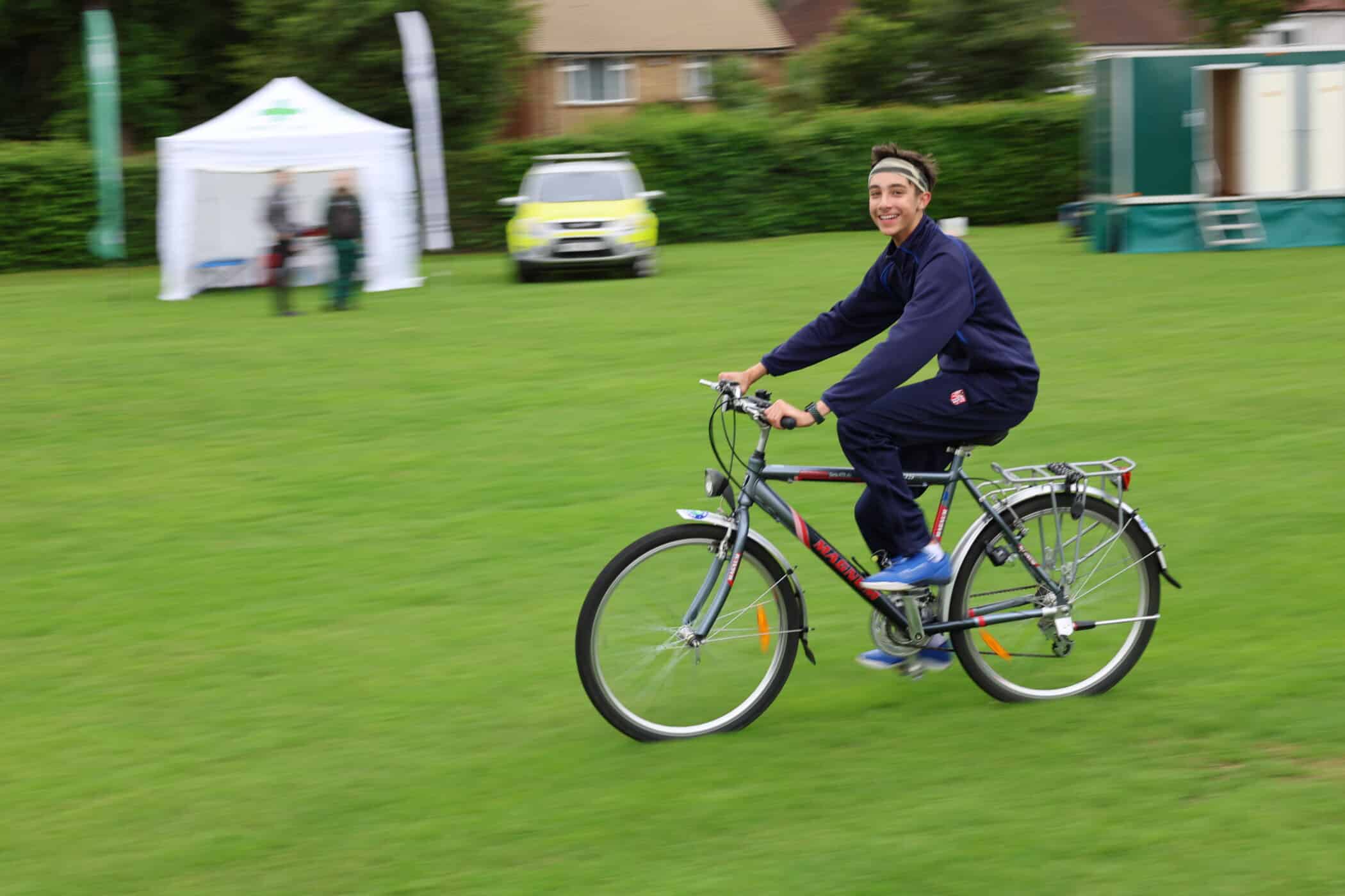 A person in sportswear rides a bicycle across a grassy field, smiling at the camera. In the background, there is a white marquee, part of a Brentwood School Foundation event, a green vehicle, and a couple of people standing.