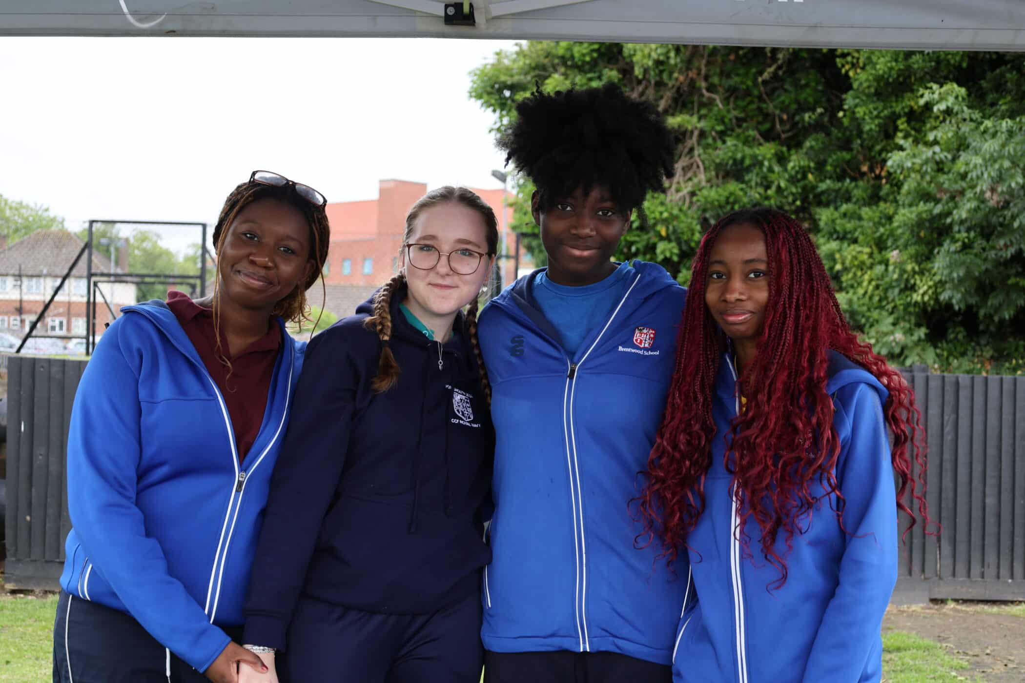 Four young women stand together outdoors, smiling at the camera. Three wear blue jackets, and one wears a dark hoodie. Trees, a fence, and buildings are visible in the background, capturing a candid moment supported by the Brentwood School Foundation.