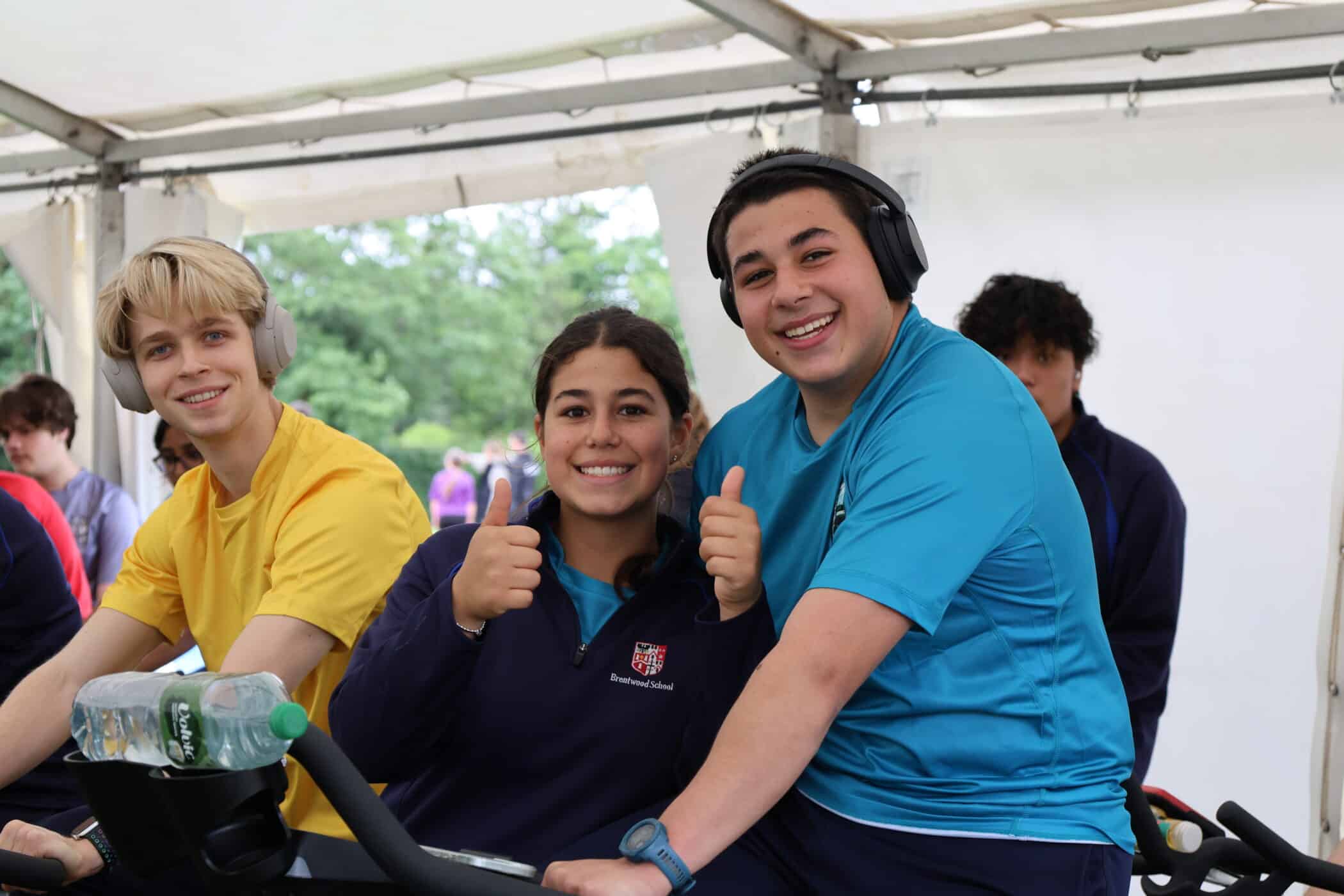 Three young people on exercise bikes smile and give thumbs up. Two of them wear headphones, and all are dressed in sports clothes under a white marquee at a Brentwood School Foundation event. Trees and more people are visible outside the marquee in the background.