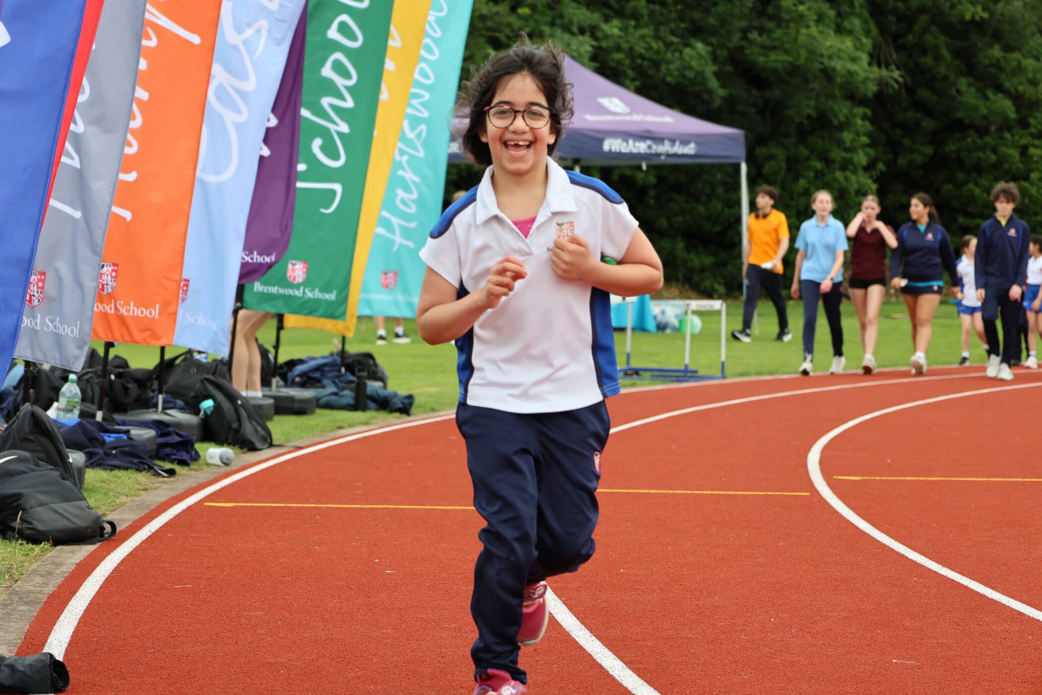 A smiling child in sports clothes runs on a red track, representing the spirit of the Brentwood School Foundation, with colourful school flags and other pupils standing in the background near a green, wooded area.