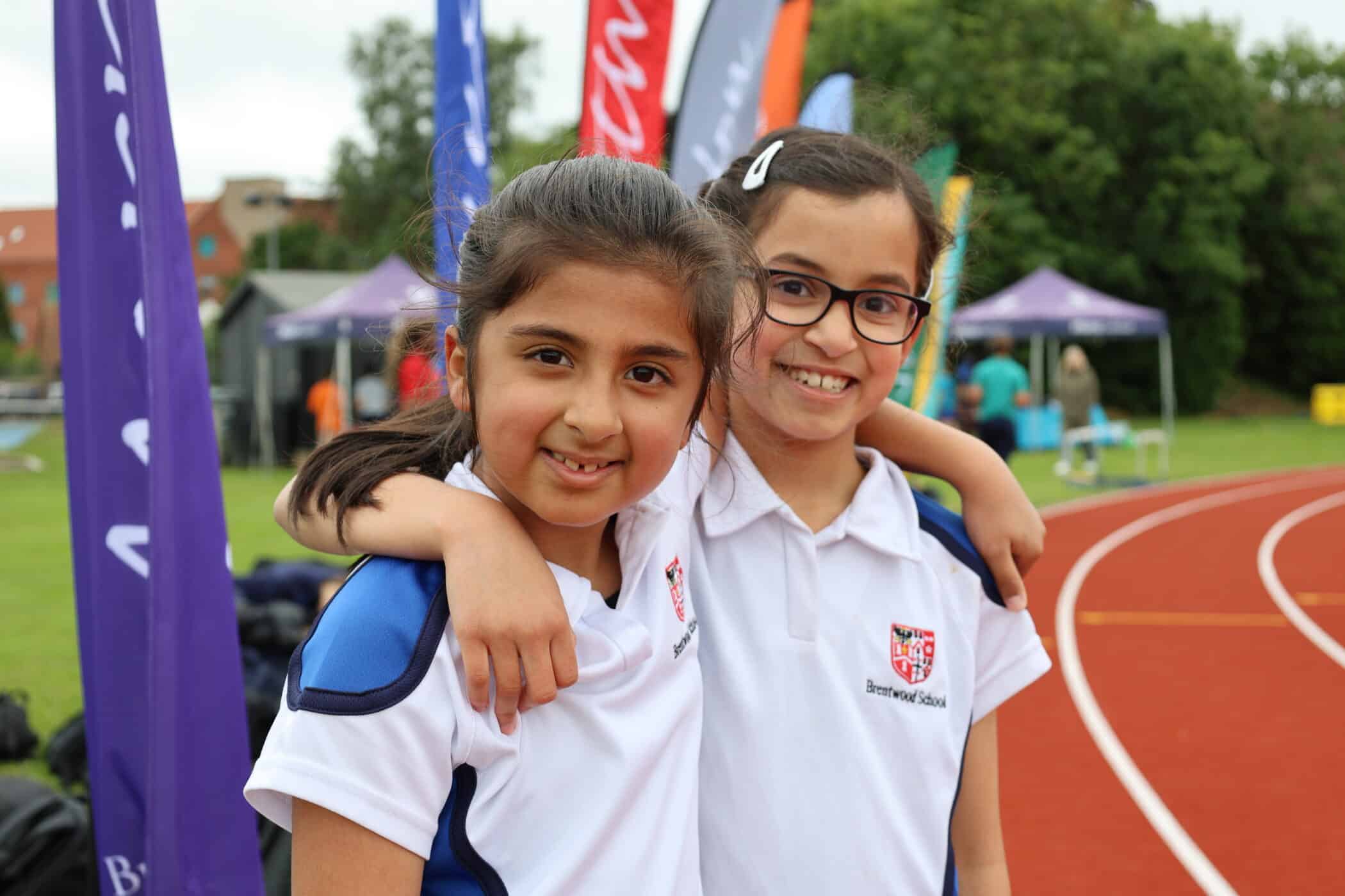 Two girls in sports kits smiling with arms round each other, standing on a running track with colourful banners and tents in the background, celebrate their achievements at a Brentwood School Foundation event.