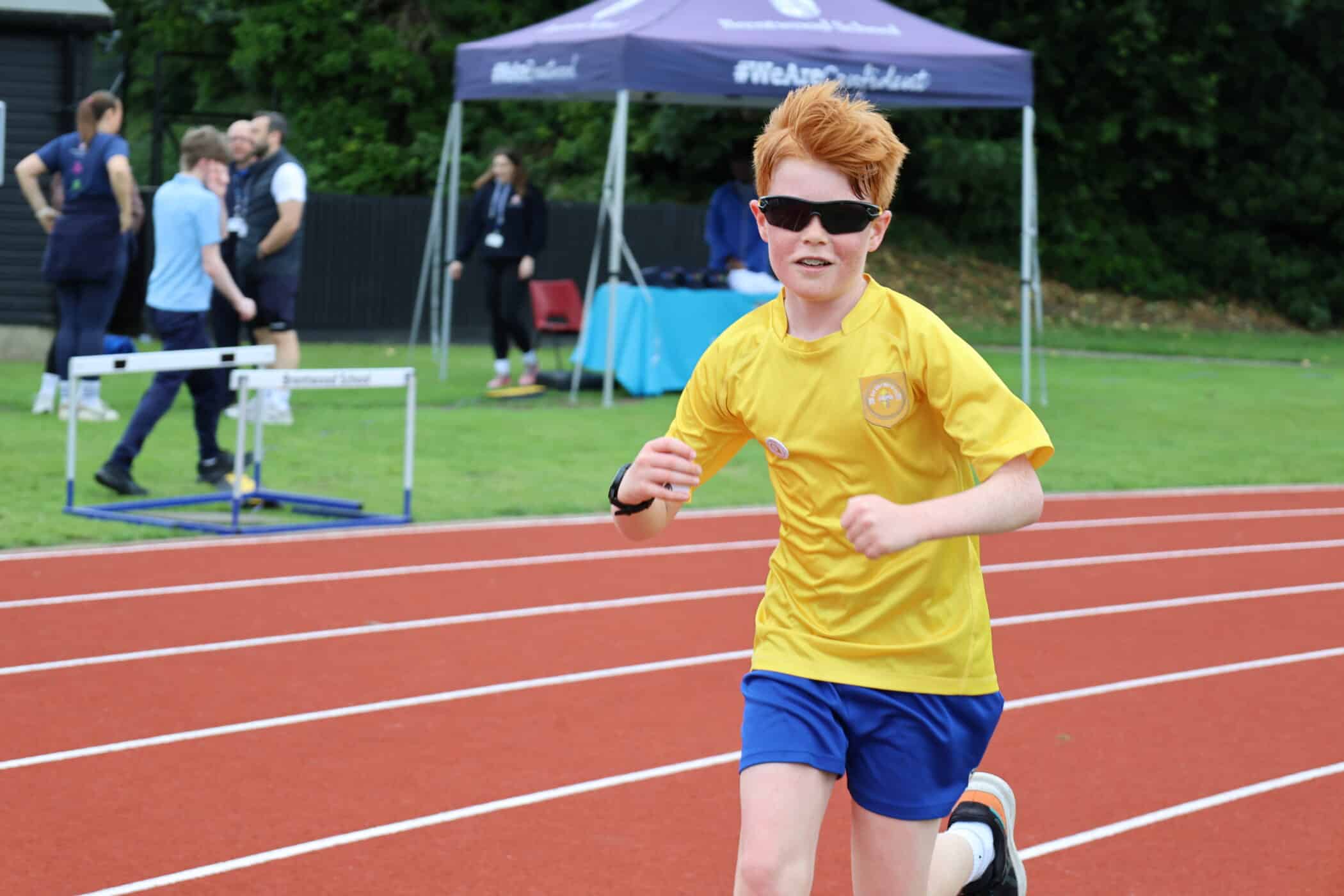 A boy with red hair and sunglasses runs on a track wearing a yellow shirt and blue shorts at the Brentwood School Foundation event. People and a hurdle are visible in the background near a marquee.