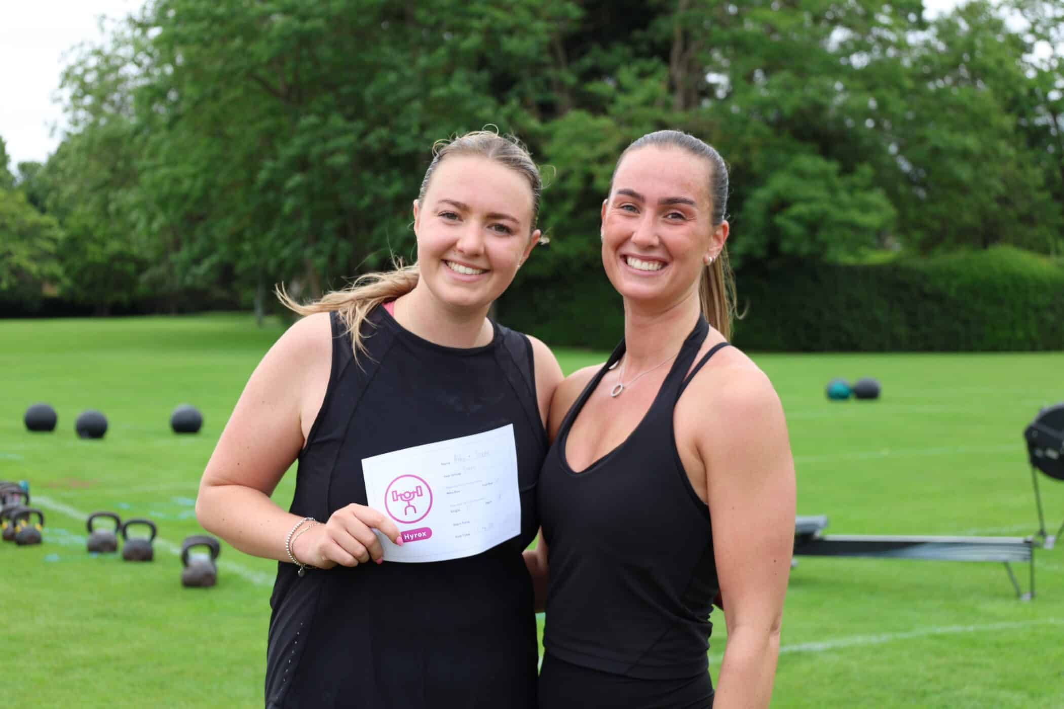 Two women in sportswear stand smiling on a grassy field. One holds a sheet of paper with writing and a pink Brentwood School Foundation logo. Exercise equipment is scattered on the grass behind them, with trees in the background.