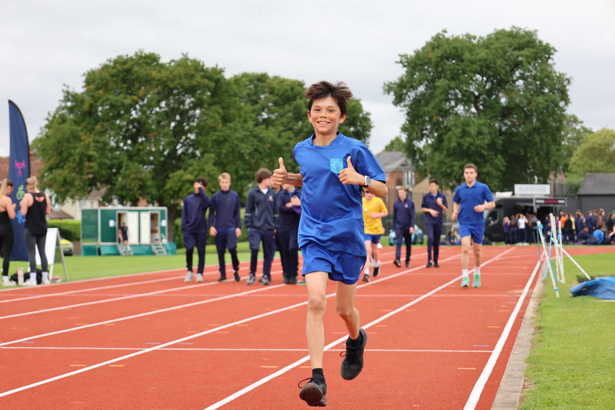 A boy in a blue outfit runs on a red track, smiling and giving a thumbs up, at an event supported by the Brentwood School Foundation. Other people are walking or standing in the background, with trees and outdoor sports facilities visible.