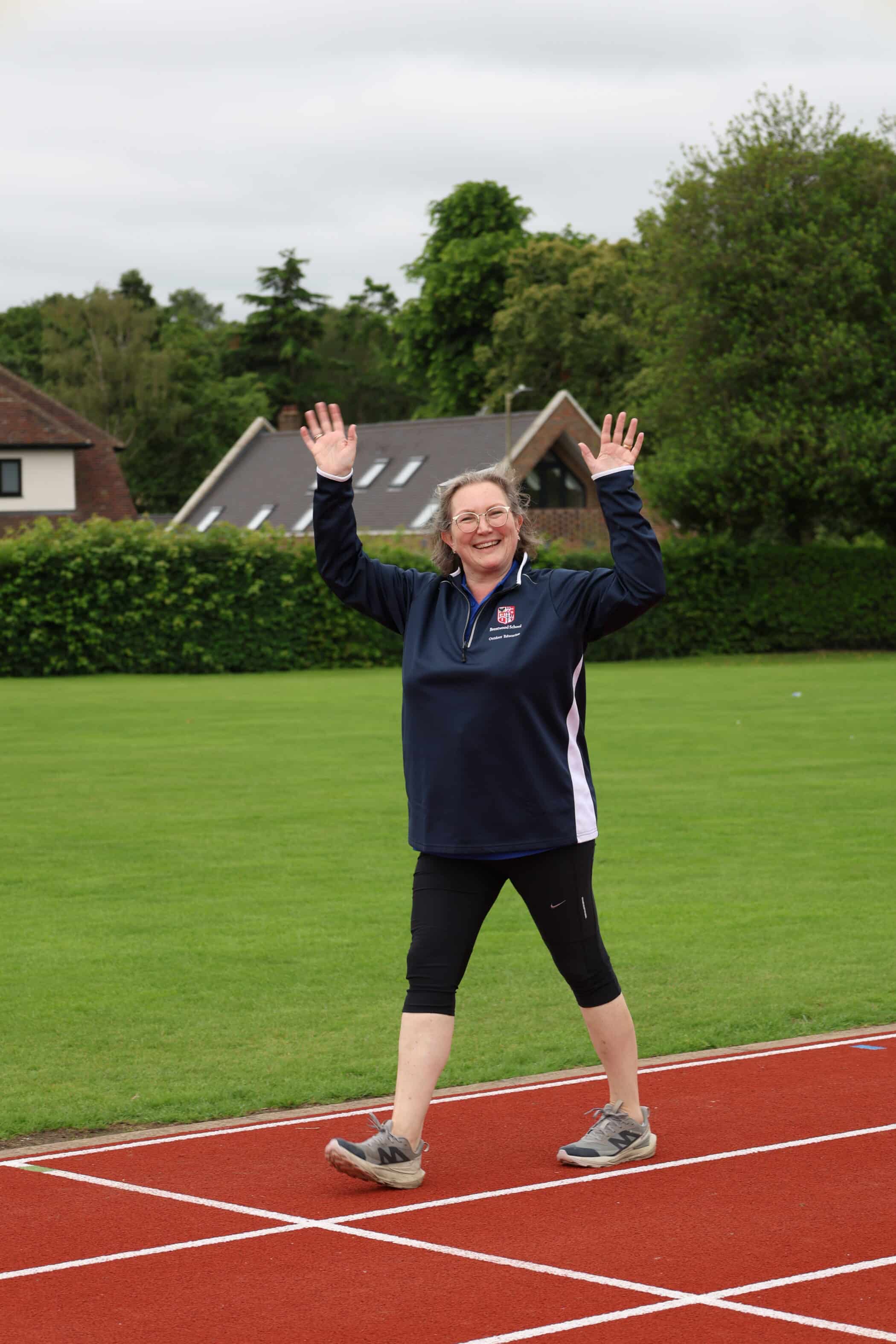 A smiling woman in sportswear walks on an athletics track with her hands raised, outdoors on a cloudy day with green grass and houses in the background, celebrating after participating in an event organised by the Brentwood School Foundation.