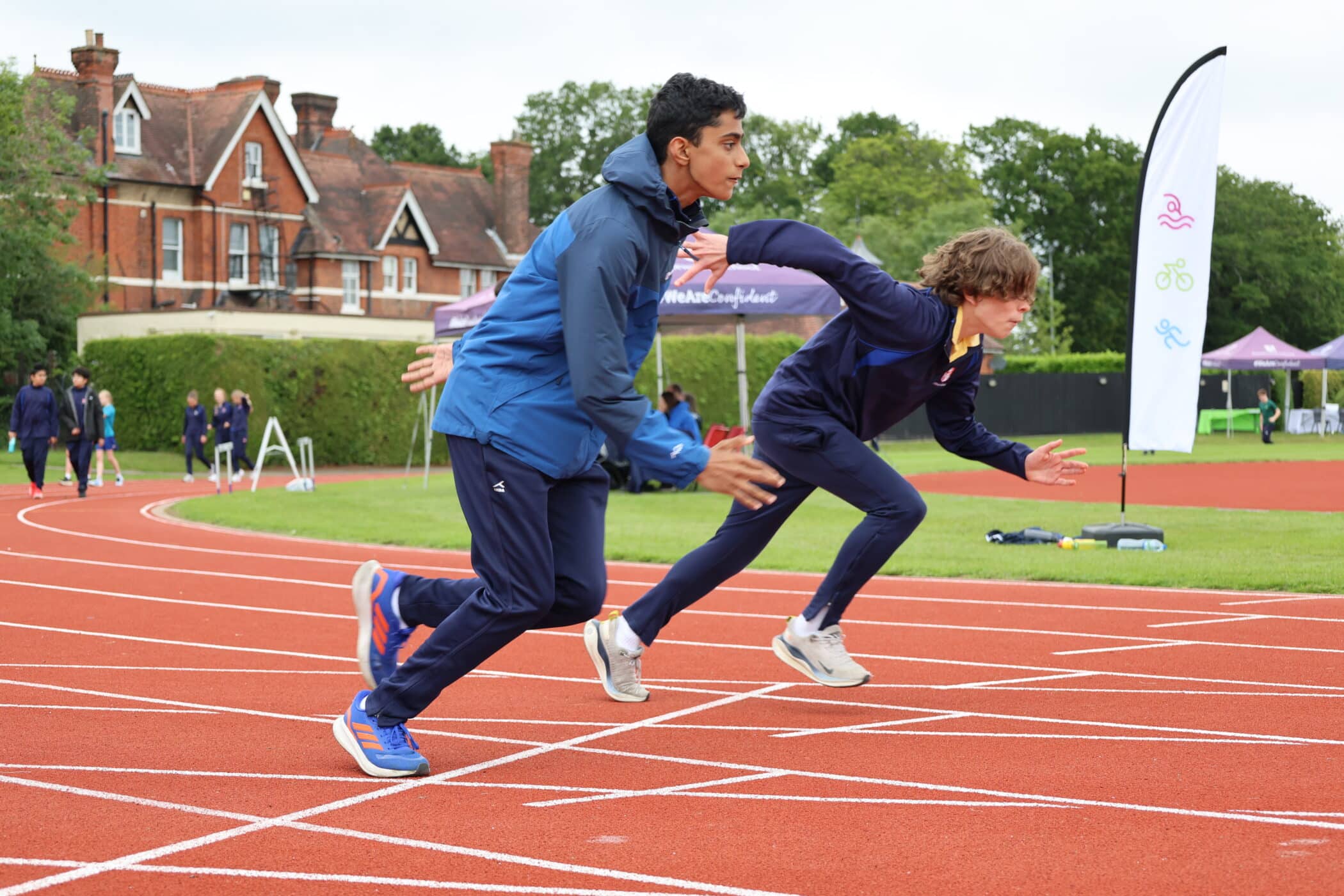 Two teenage boys in blue jackets sprint from the starting line on a red outdoor track, with buildings, trees, and event banners, proudly displaying the Brentwood School Foundation in the background.