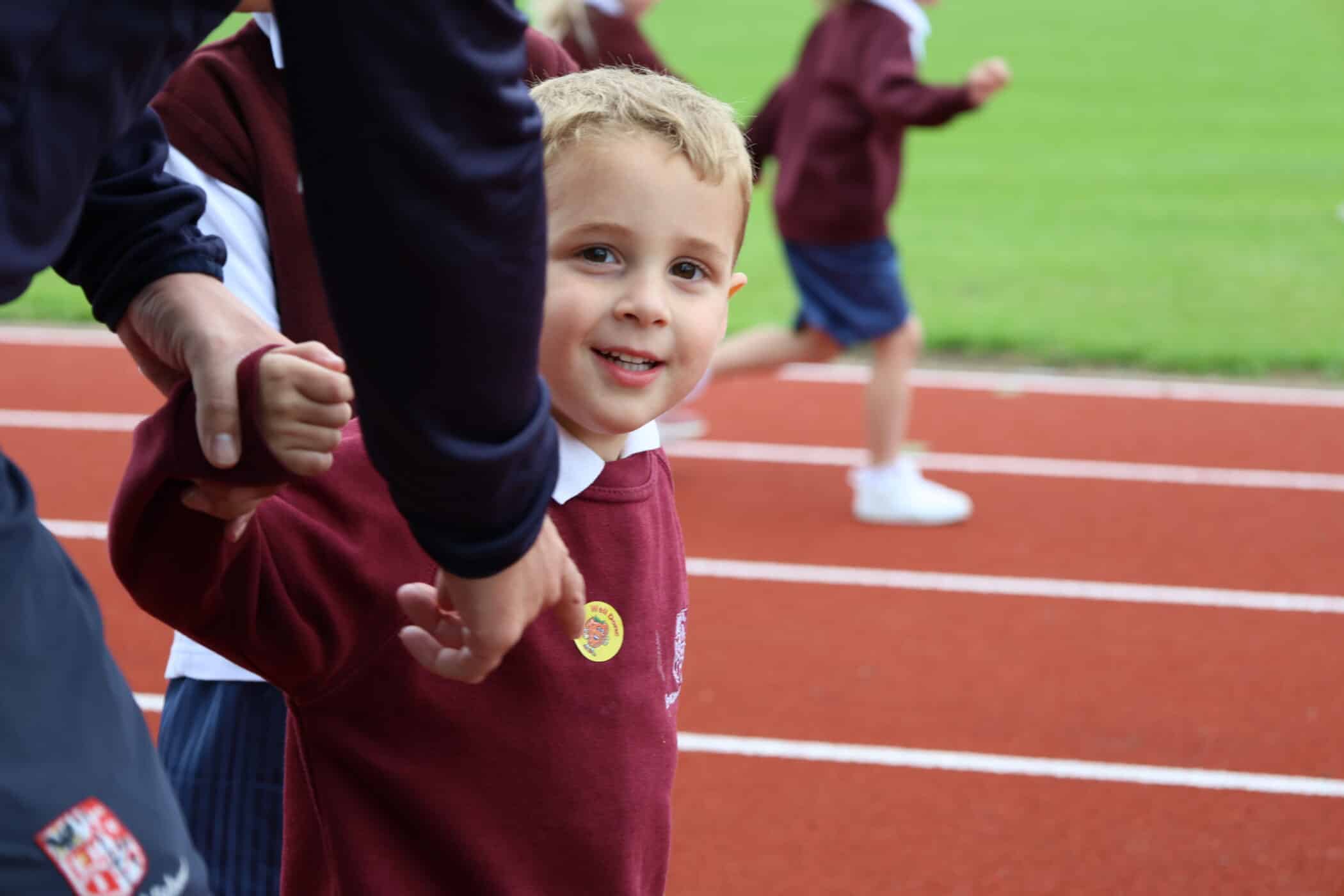 A young boy in a maroon jumper smiles whilst holding an adult’s hand on a running track, his joy reflecting the supportive environment fostered by the Brentwood School Foundation. Other children run in the background.