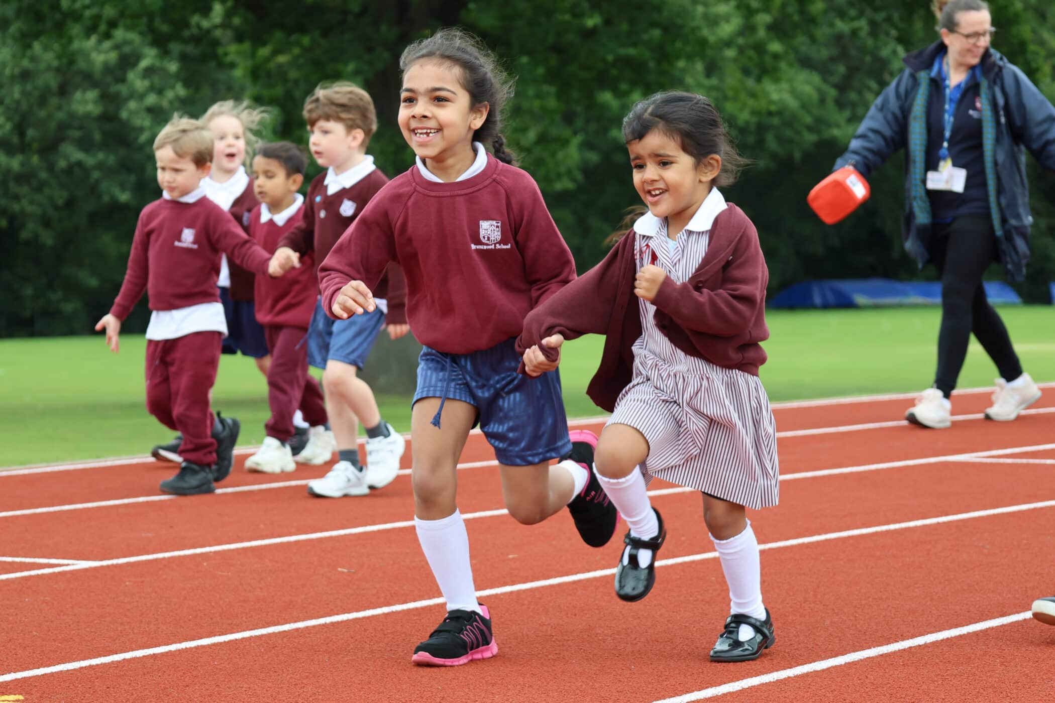 Two young girls hold hands and run on a track, smiling, with other children and an adult in the background. All the children wear school uniforms and are outdoors near greenery, reflecting the joyful spirit fostered by the Brentwood School Foundation.