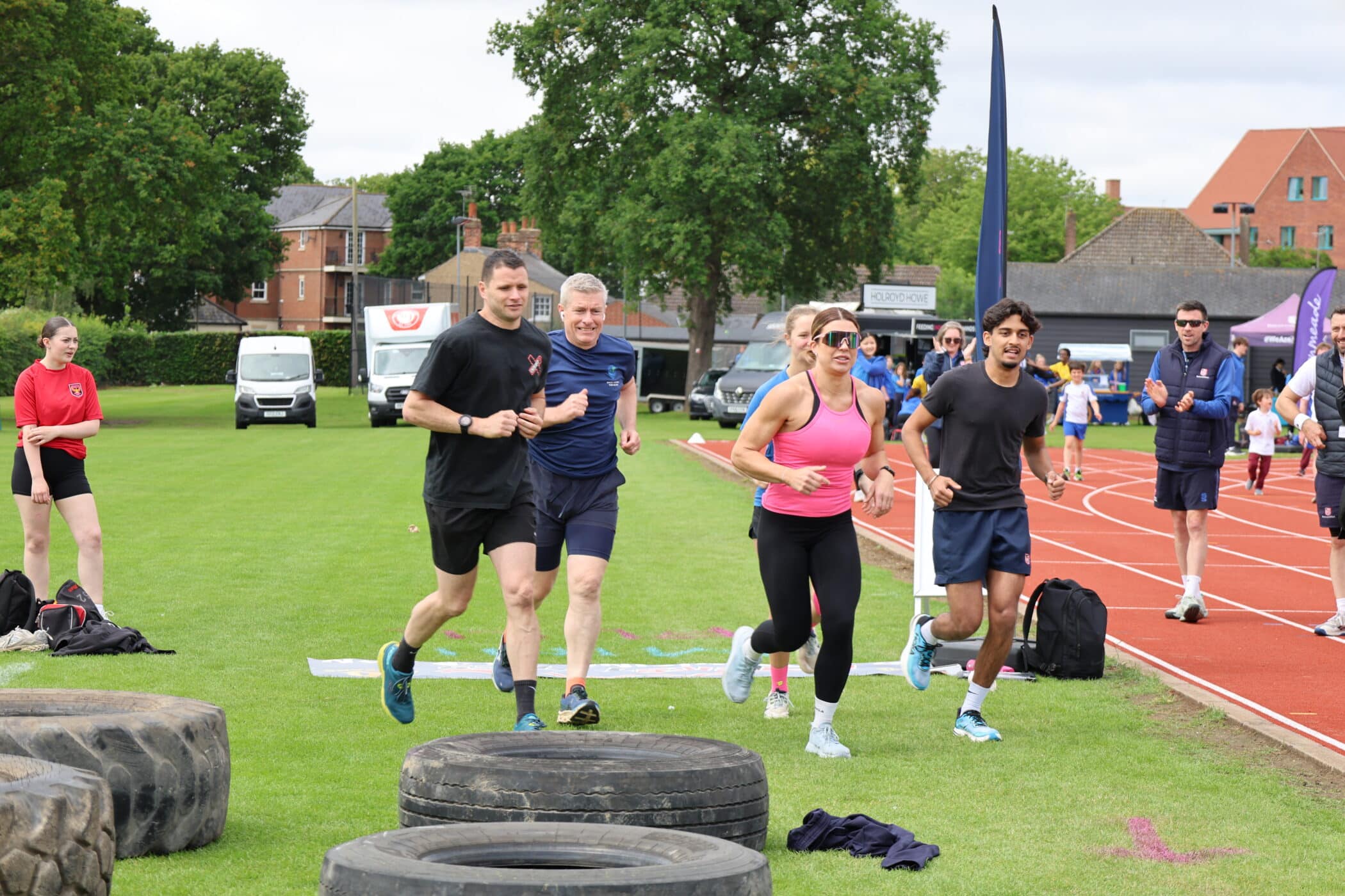 Four people are jogging together on grass beside a running track, with large tyres on the ground in front of them, participating in an activity organised by the Brentwood School Foundation. Other people and buildings are visible in the background.