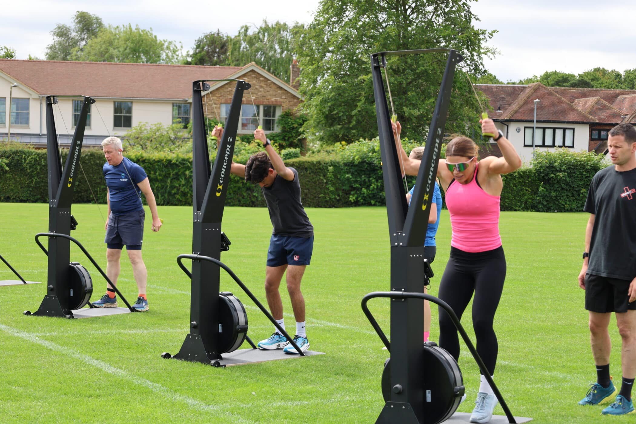 Four people exercising outdoors on ski erg machines on a grassy field, with buildings and trees in the background. Two trainers from the Brentwood School Foundation observe participants who are actively pulling the ski handles.