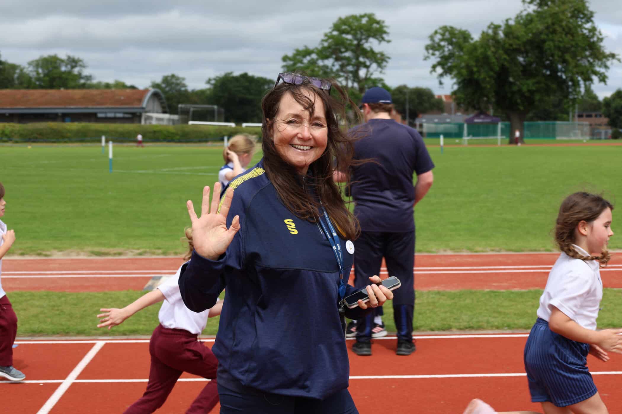 A smiling woman in a navy tracksuit waves at the camera on a running track, whilst children run in the background during an outdoor sports event organised by the Brentwood School Foundation.
