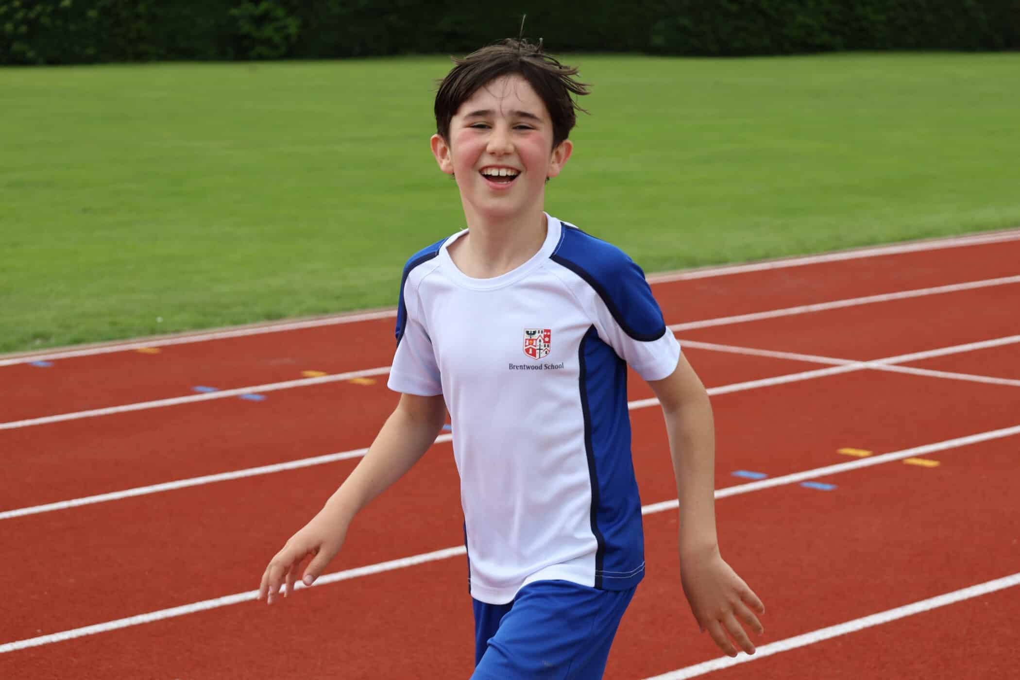 Smiling boy in a white and blue sports kit running on a red track, with green grass and shrubs in the background, embodying the spirit of the Brentwood School Foundation.