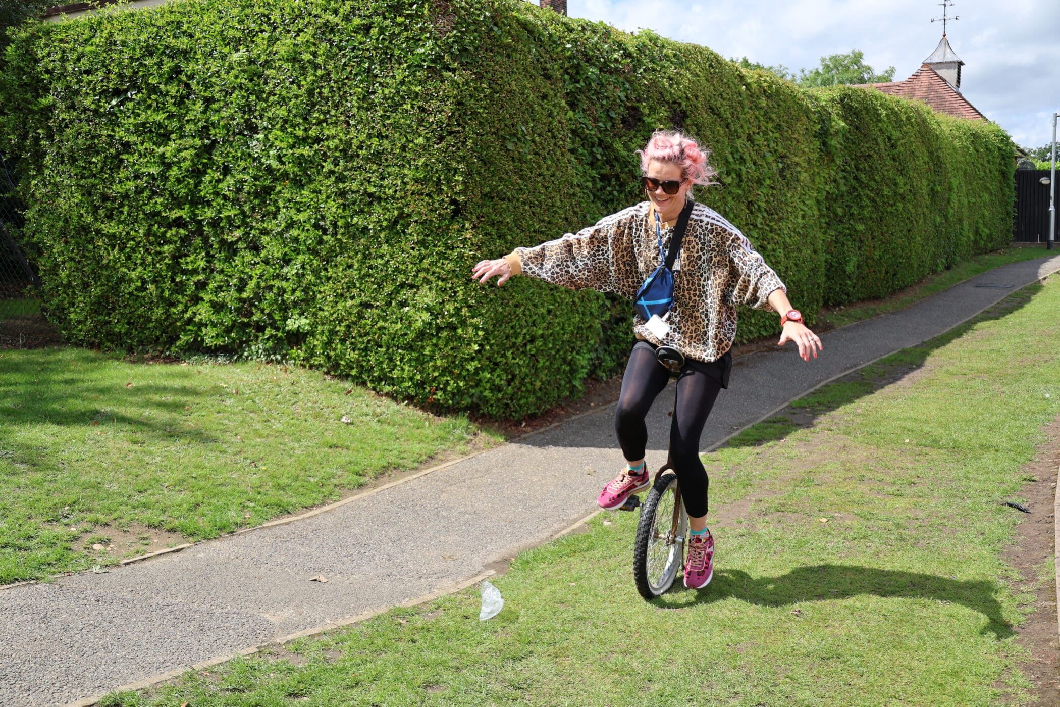 A person with pink hair, sunglasses, and a leopard-print jacket rides a unicycle on grass next to a path, arms outstretched for balance, a scene that captures the vibrant spirit often celebrated by the Brentwood School Foundation. There’s a tall green hedge and cloudy sky in the background.