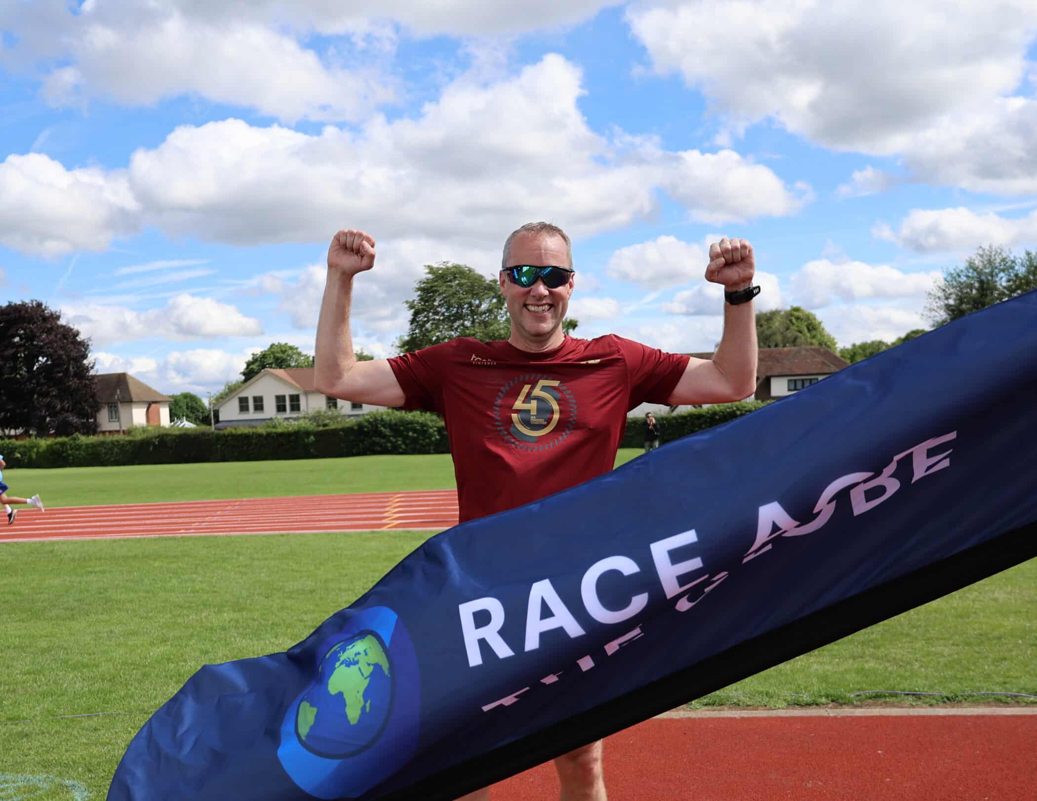 A smiling runner in a red shirt raises his arms in victory as he crosses the finish line at an outdoor track, with a Race Across The Globe banner in front, an inspiring moment supported by the Brentwood School Foundation and houses and trees in the background.