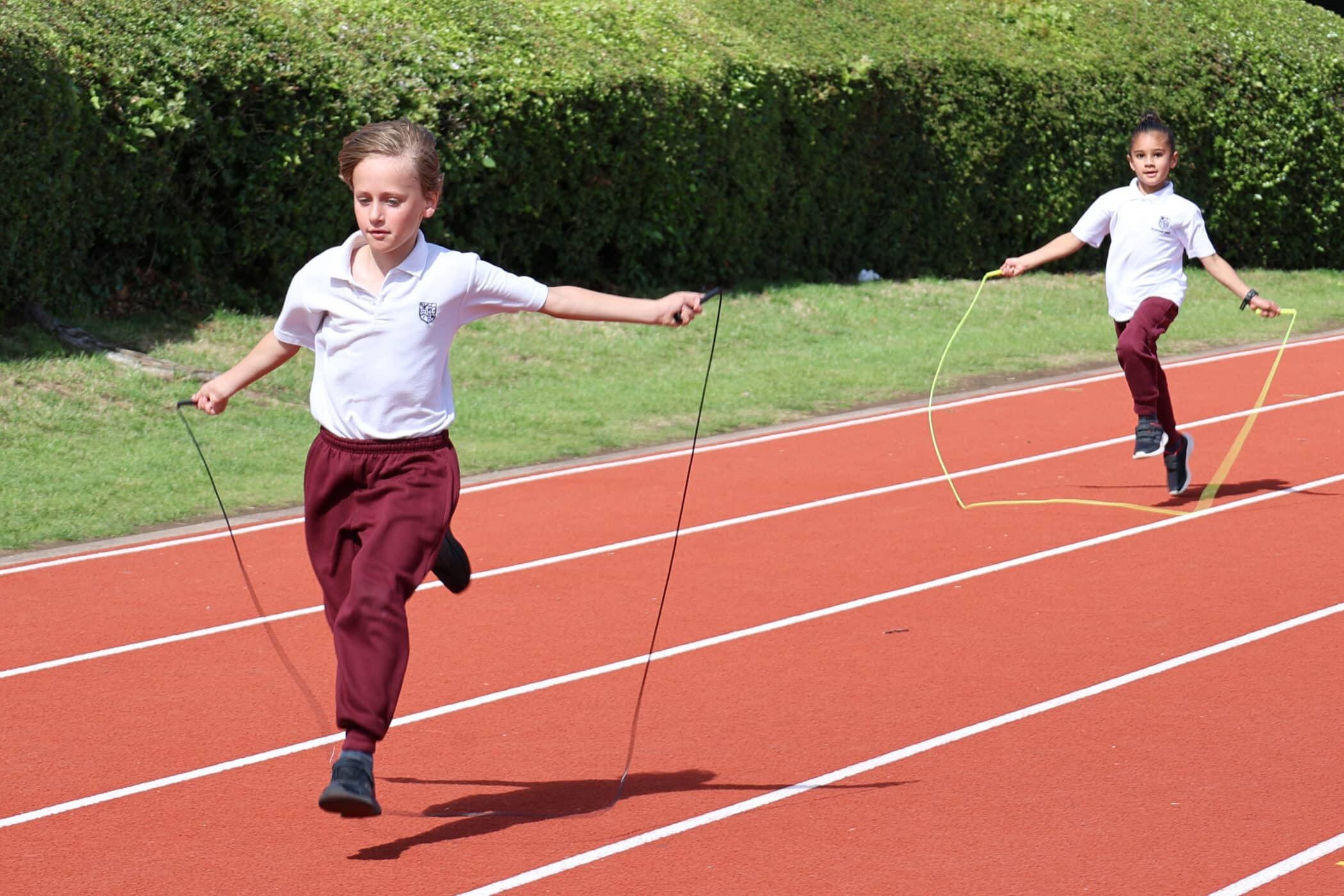 Two children in white shirts and maroon trousers are skipping rope on a red running track outside, with green bushes in the background, a vibrant scene supported by the Brentwood School Foundation.