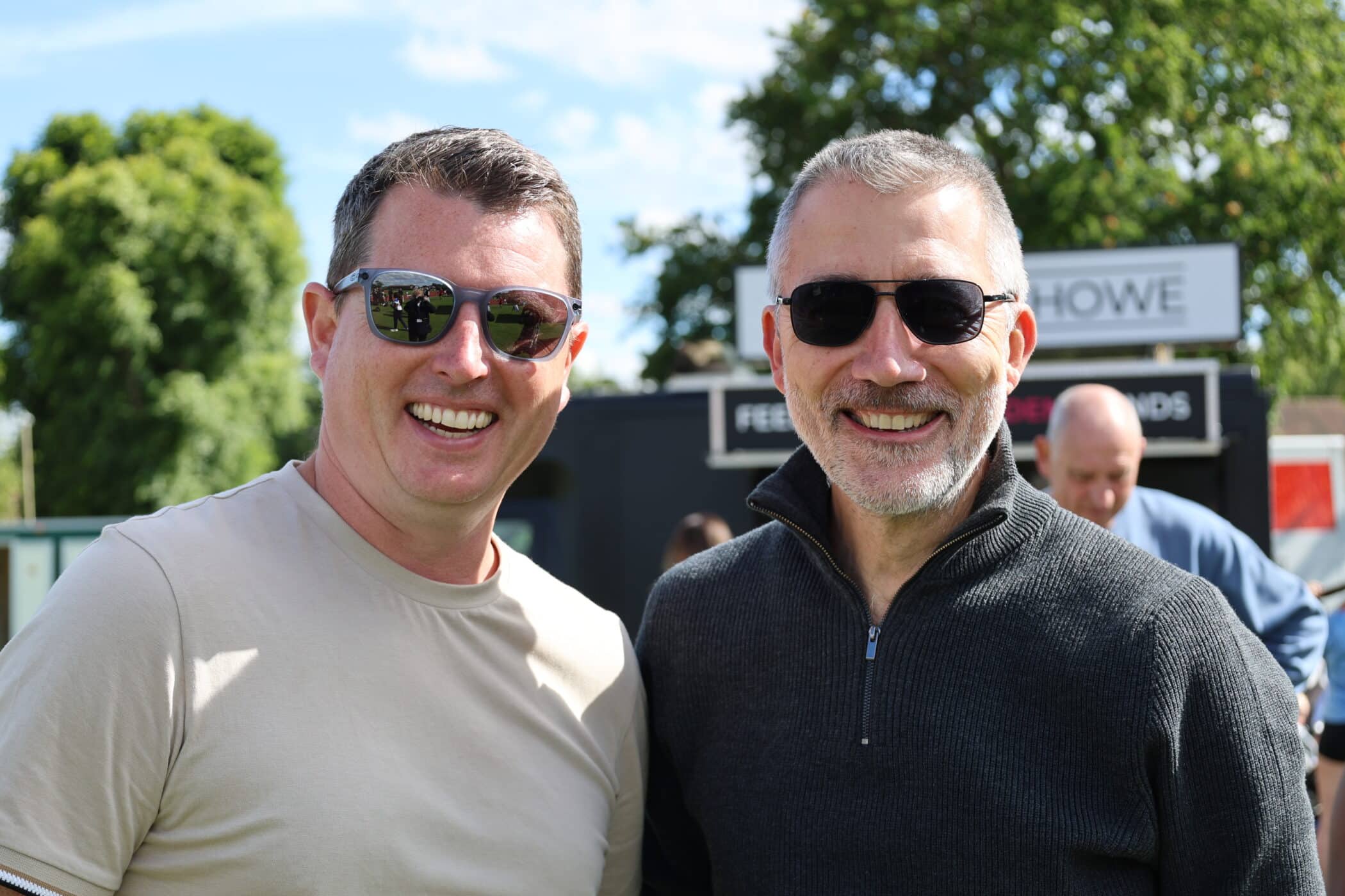 Two smiling men wearing sunglasses stand outdoors on a sunny day at a Brentwood School Foundation event. Both are casually dressed and appear to be enjoying the occasion.