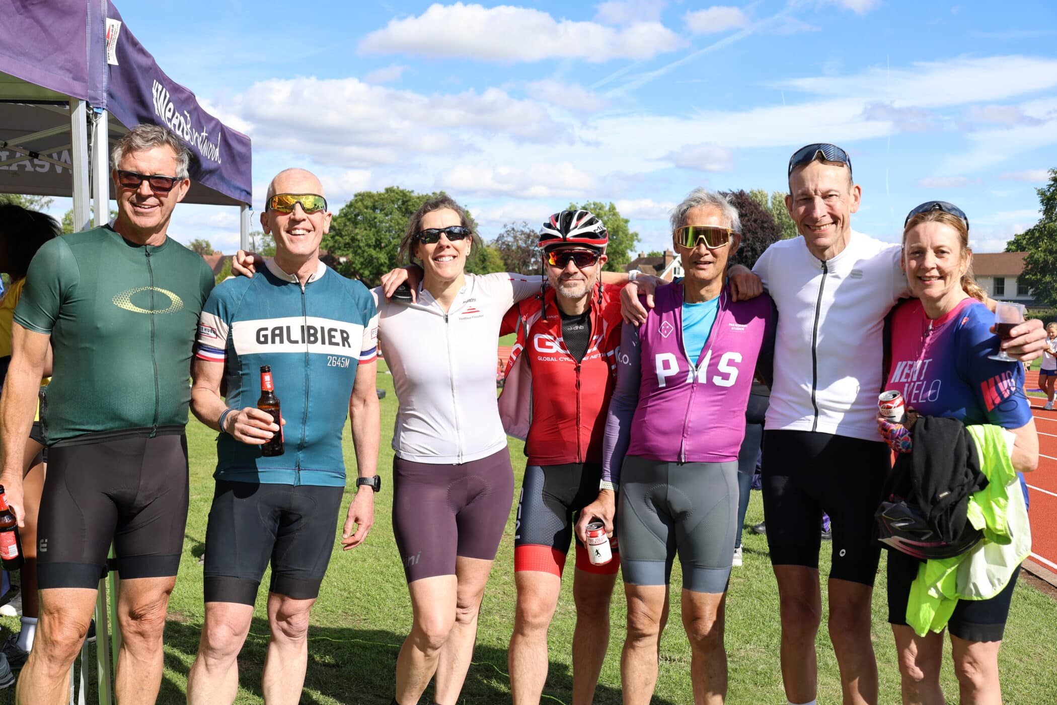 Seven cyclists in colourful gear stand together smiling, holding drinks, on a sunny outdoor field with blue sky and clouds. Some wear sunglasses and helmets, and a tent is visible in the background.