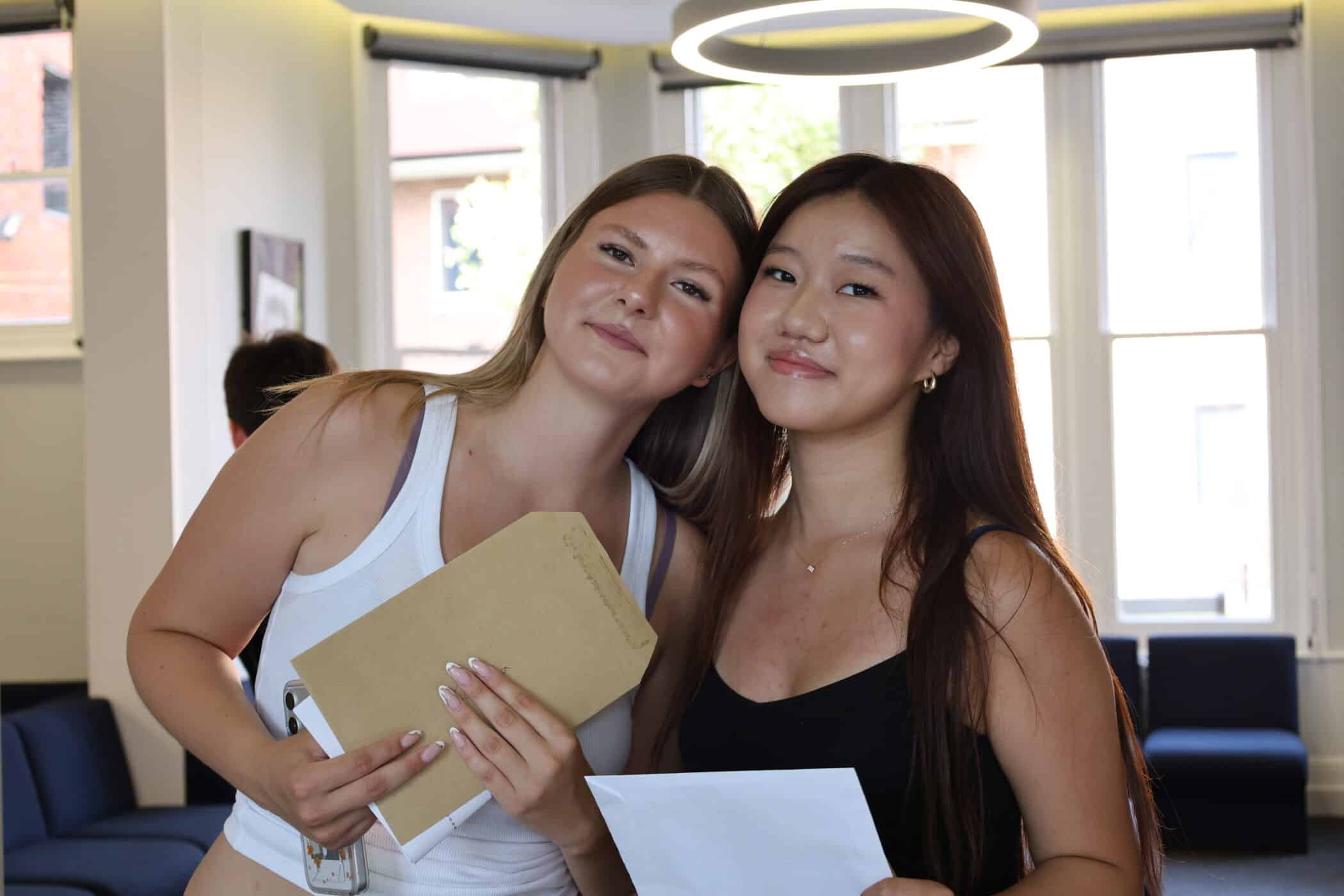 Two young women standing indoors, smiling at the camera. One holds a brown envelope and the other holds a sheet of paper. Bright window light fills the room behind them.