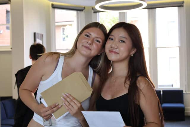 Two young women smiling and standing close together indoors, one holding a brown envelope and the other holding a sheet of paper, with large windows and modern décor in the background.