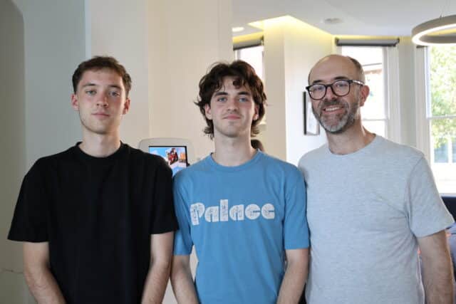 Three men stand indoors, posing for a photo. Two younger men are on the left, one wearing a black shirt and the other a blue “Palace” shirt. An older man with glasses and a beard stands on the right in a light grey shirt.