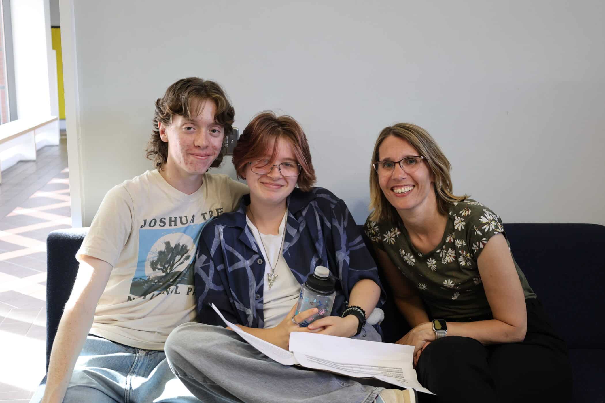 Three people sit closely together on a sofa, smiling at the camera. Two young people are holding papers and a water bottle, and an adult woman sits on the right, wearing glasses. The background is a plain wall with light from a window.