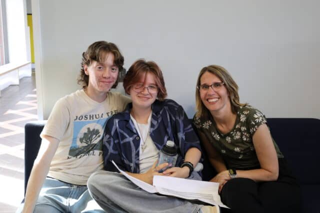Three people sit closely together on a sofa, smiling at the camera. Two young people are holding papers and a water bottle, and an adult woman sits on the right, wearing glasses. The background is a plain wall with light from a window.