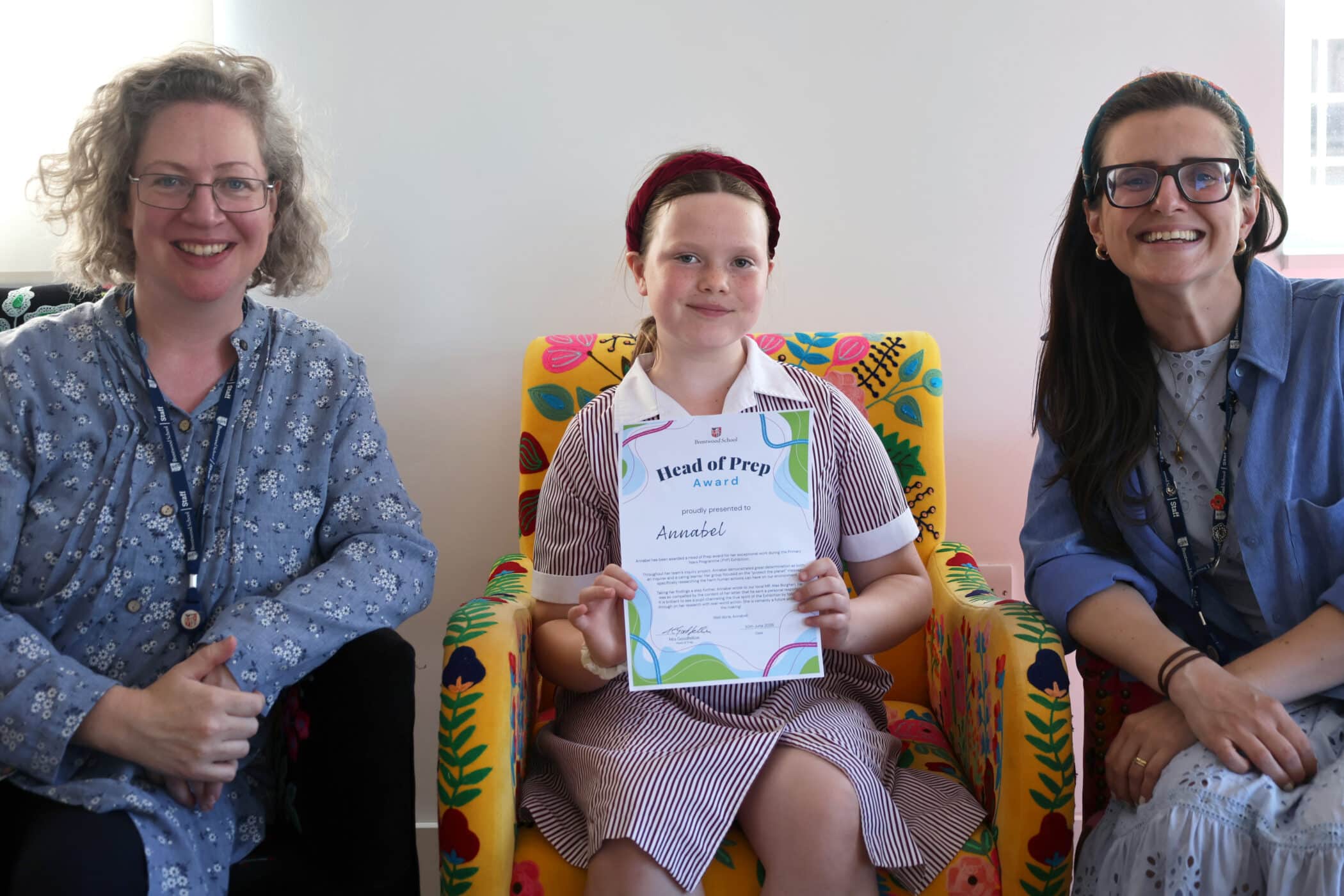 A girl in a school uniform sits on a colourful chair, holding an award certificate and smiling, with two women in glasses and patterned shirts sitting on either side of her.