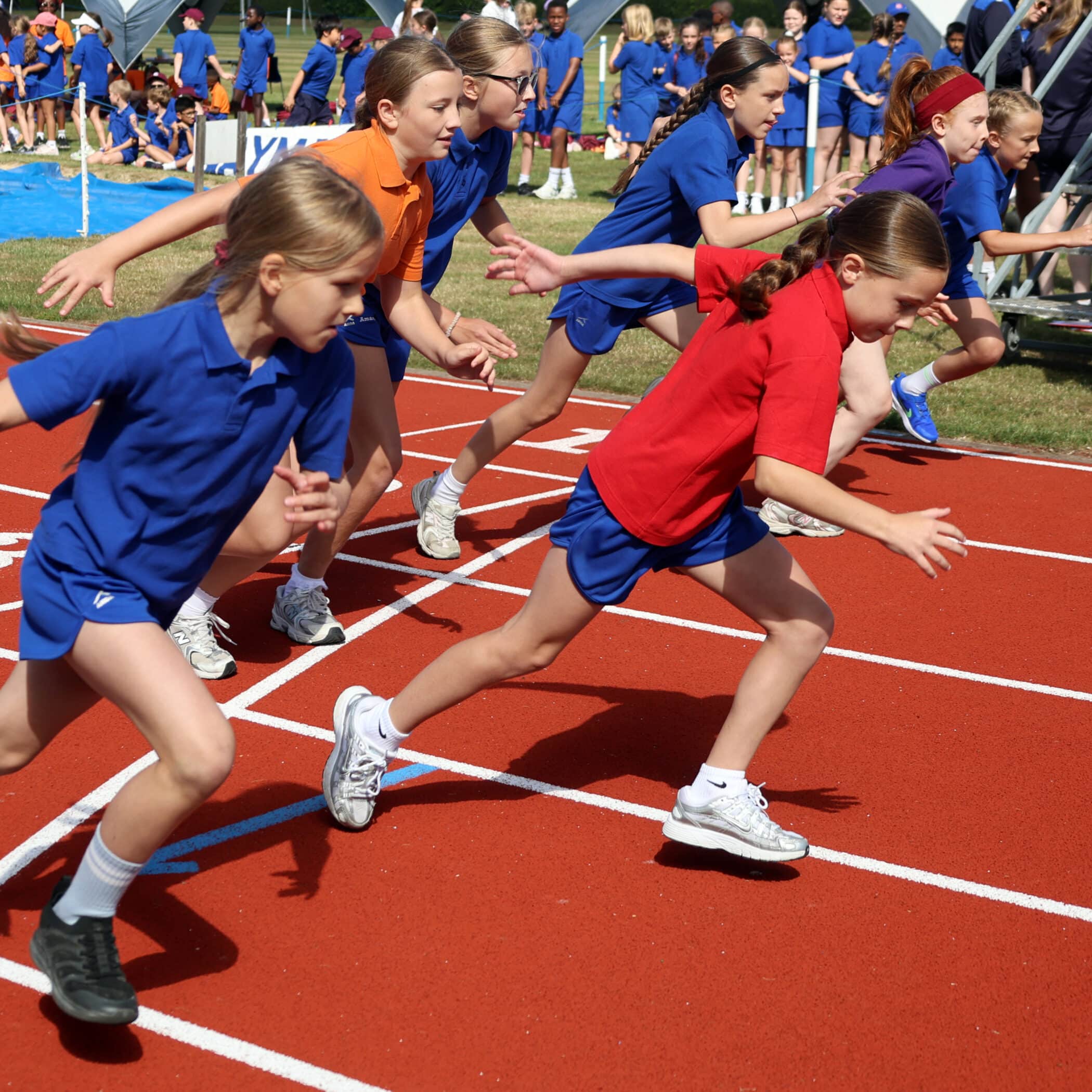 A group of young girls in colourful sports kit sprint off the starting line in a race on a red running track, with spectators and other participants in the background.