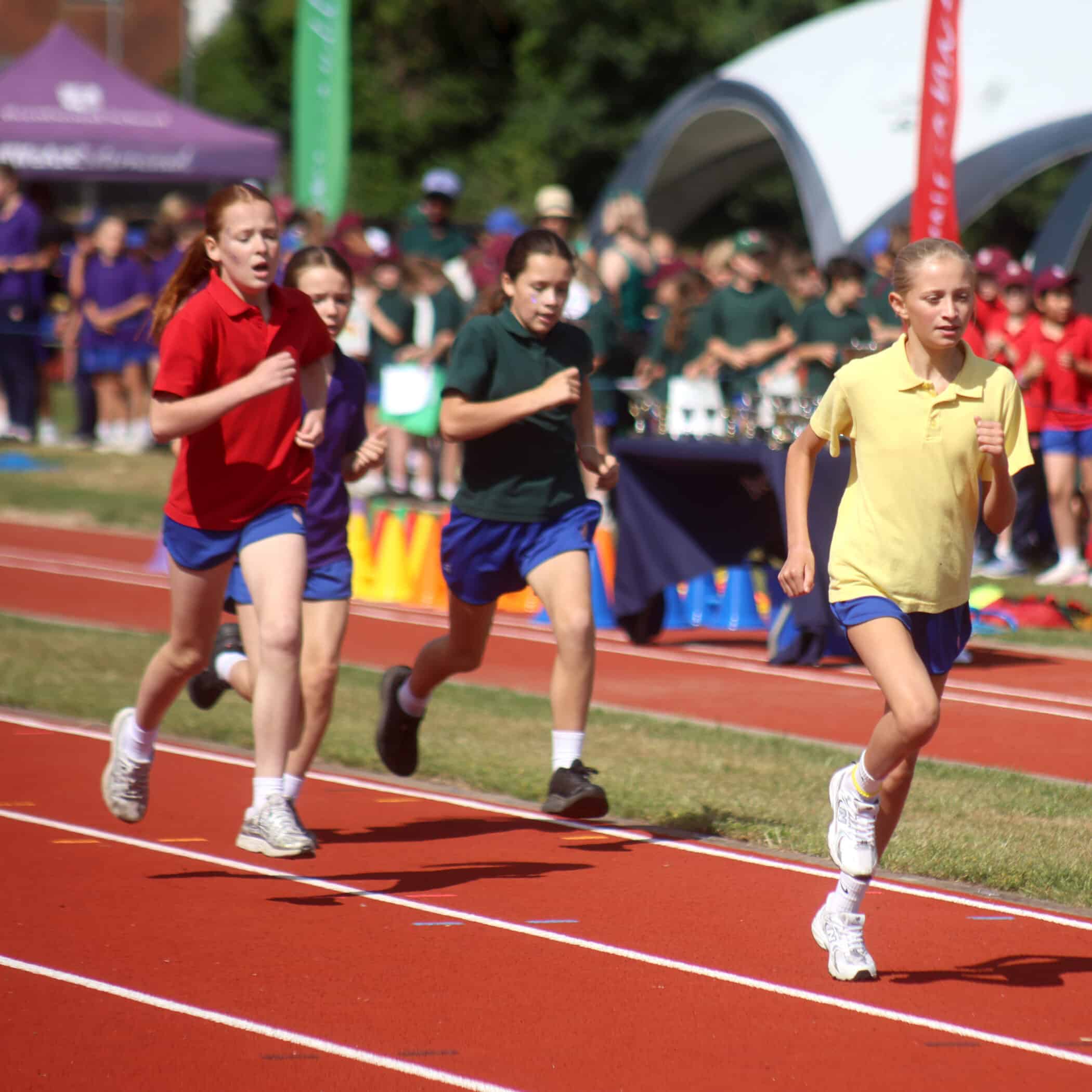 Four girls are running on a red track during a school sports event, each wearing differently coloured shirts and blue shorts. Spectators and tents are visible in the background.
